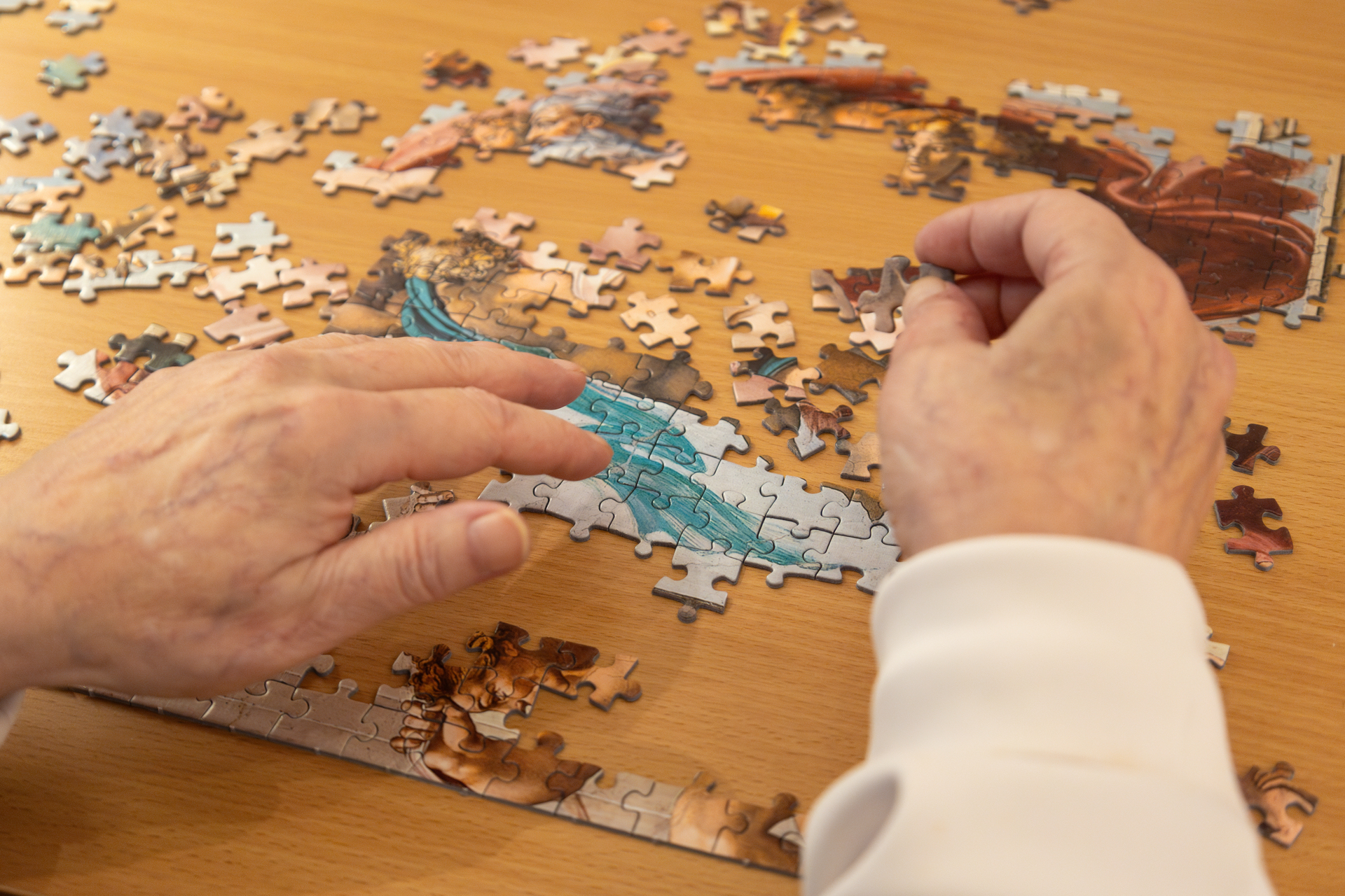 To bilder etter hverandre, der det første viser en kvinne i 60-årene med mørkt hår og briller. PÅ bordet foran henne ligger et mange brikker i et stort puslespill. Det andre er et nærbilde av hendene hennes som pusler.