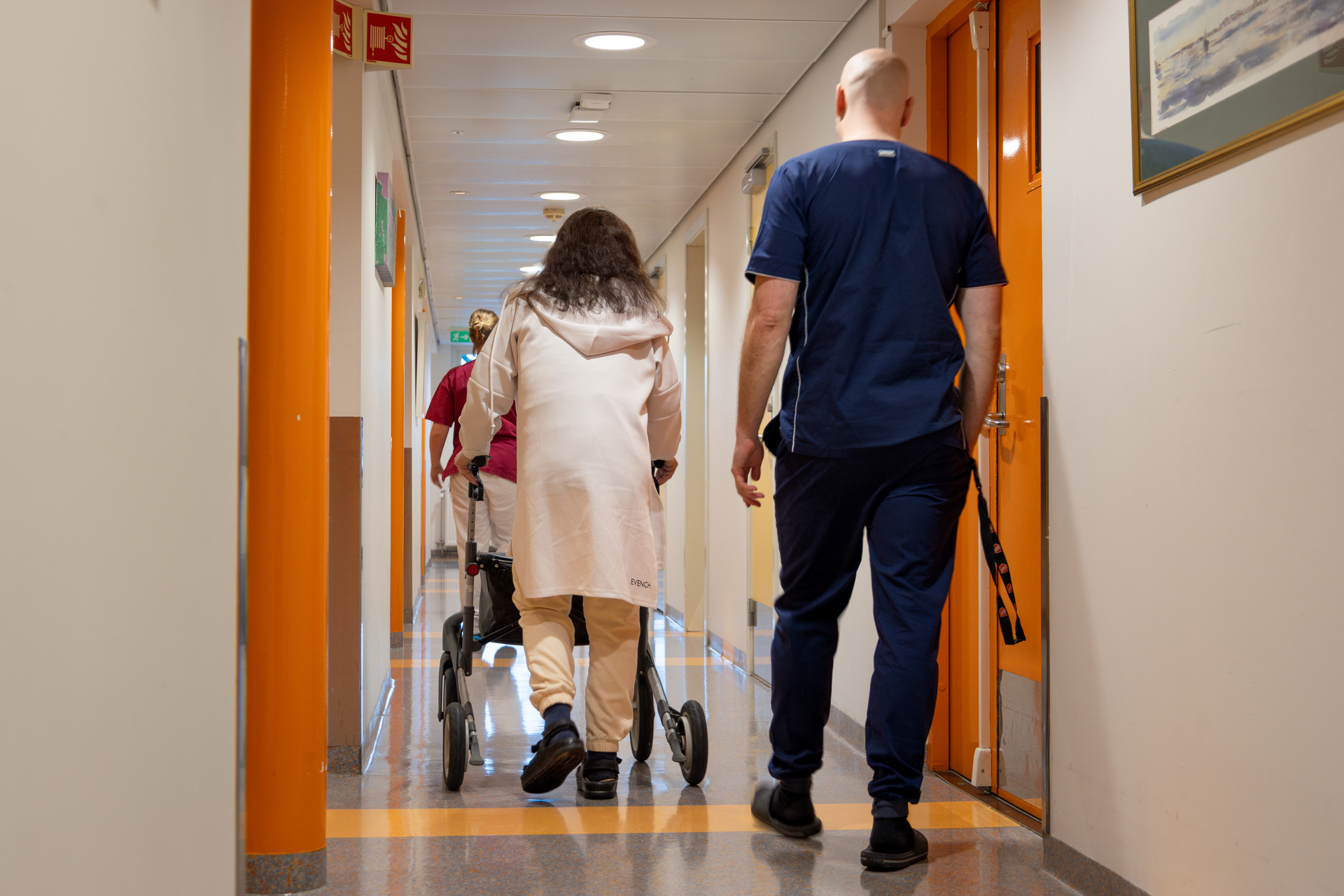Foto: Kvinne med rullator følges av mannlig sykepleier i blå uniform nedover en korridor. Vi ser ryggen på begge.