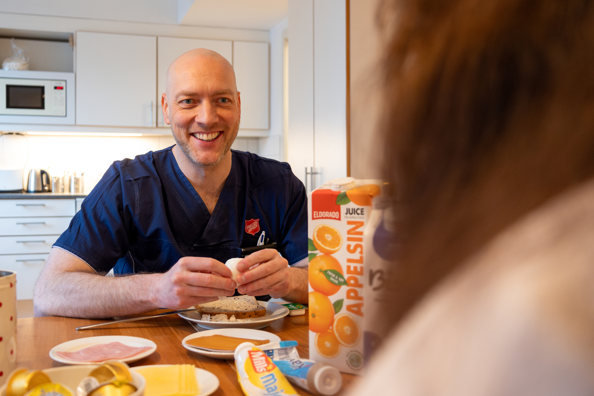 Foto: To bilder etter hverandre, der det første viser en smilende mannlig sykepleier smile. Han tar skallet av et egg ved frokostbordet, som er dekket med juice, brød og pålegg. Det andre er et nærbilde av egget som er skåret opp i biter på en brødskive.