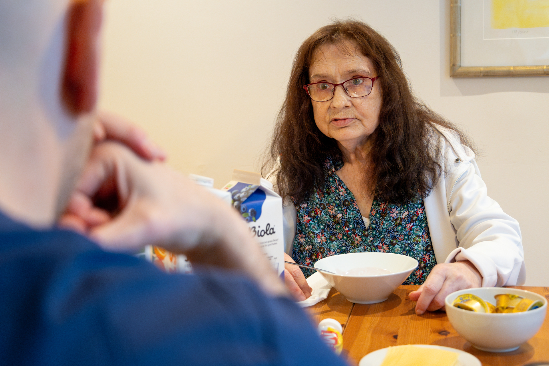 Foto: Kvinne med mørkt hår og briller sitter på den andre siden av frokostbordet. En mannlig sykeplerier i blå drakt sitter med ryggen til.