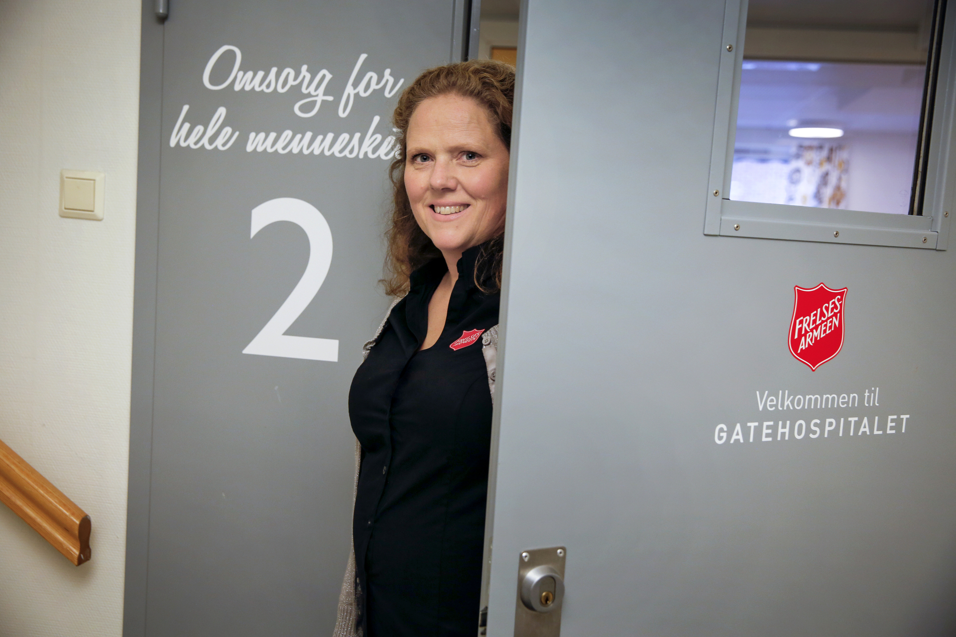 Foto: Kvinne i blå sykepleieruniform gløtter ut av en grå, tung metalldør inne på et sykehus.