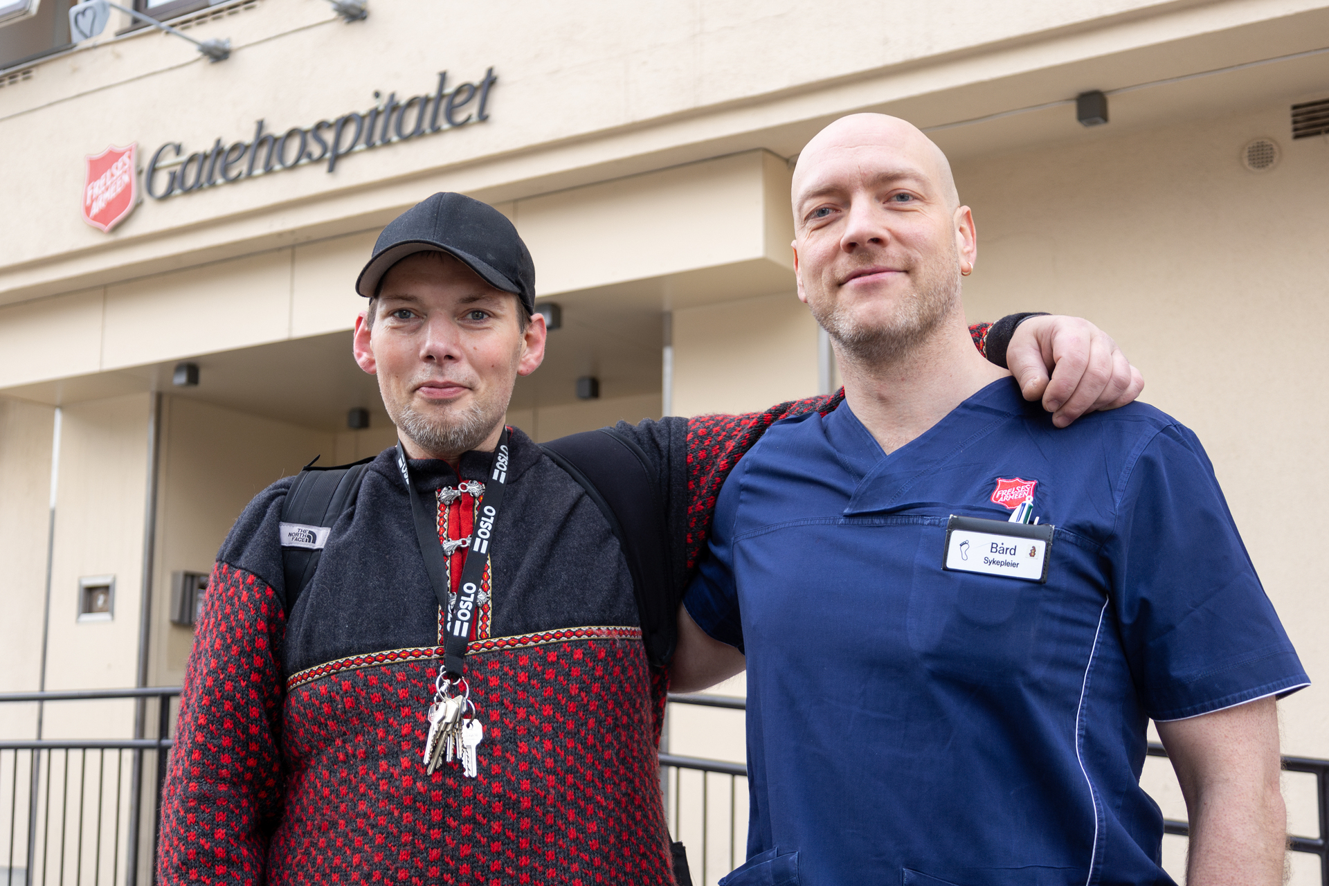Foto: To menn står utenfor inngangen til Gatehospitalet i Oslo. Han til venstre er kledd i caps og grå og rød strikkegenser. Han til høyre er iført blå sykepleierskjorte med rød logo fra Frelsesarmeen. De holder karslig om hverandre.