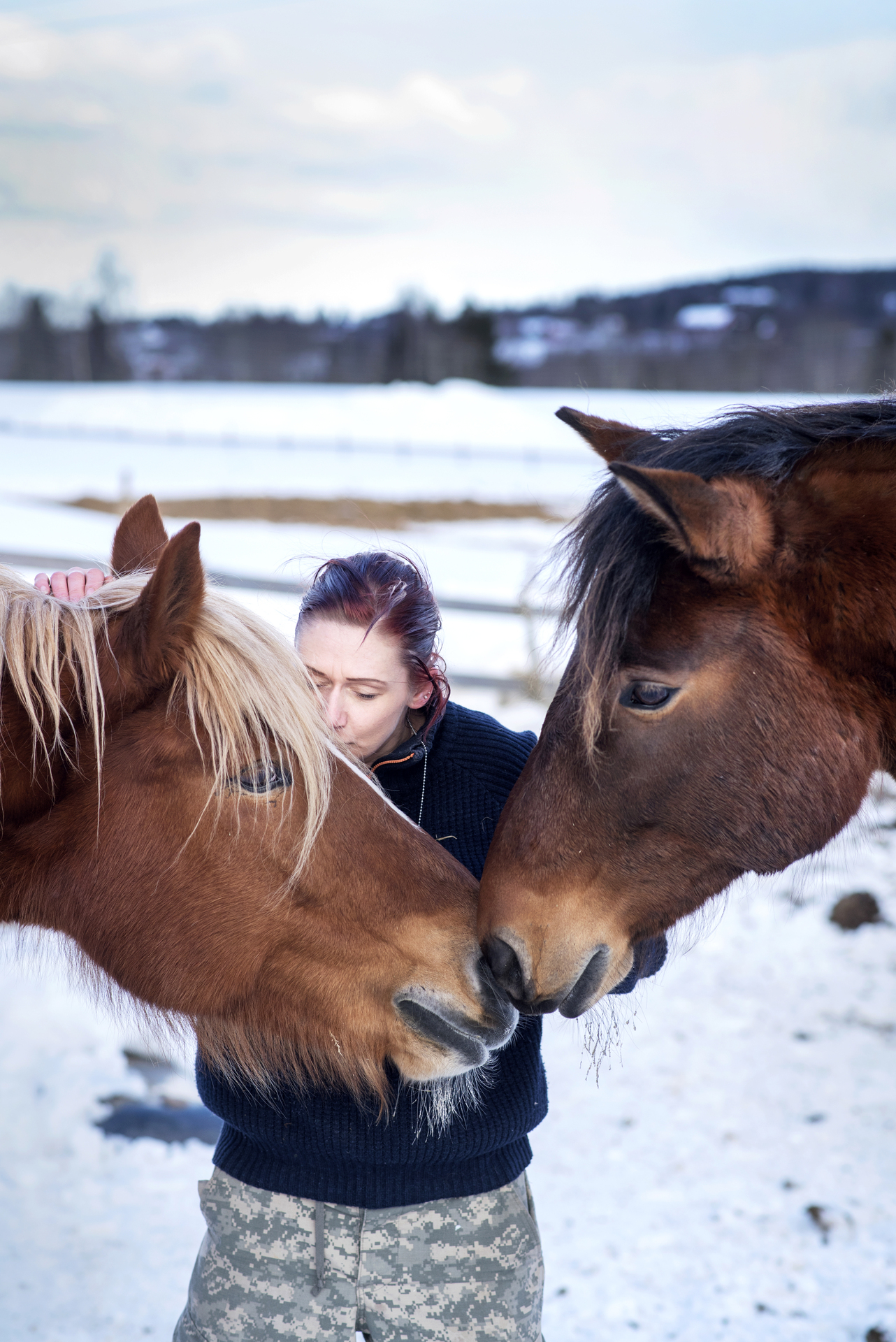 Foto: Kvinne står med lukkede øyne mellom to hester ute på en snødekket mark.
