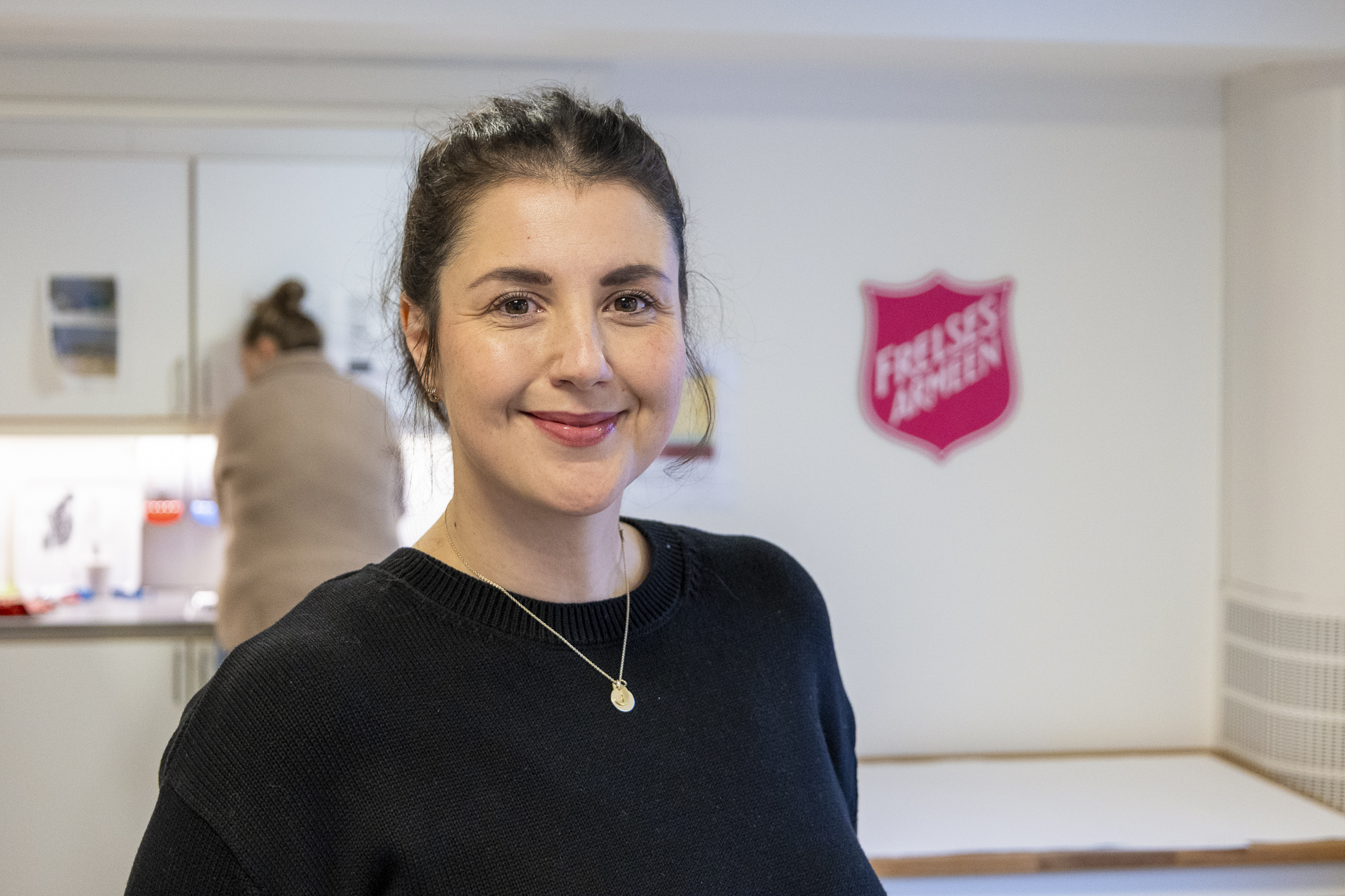 Ung kvinne står inne på et kjøkken og smiler. I bakgrunnen sees både Frelsesarmeens logo og en kvinne som ordner noe på kjøkkenbenken.