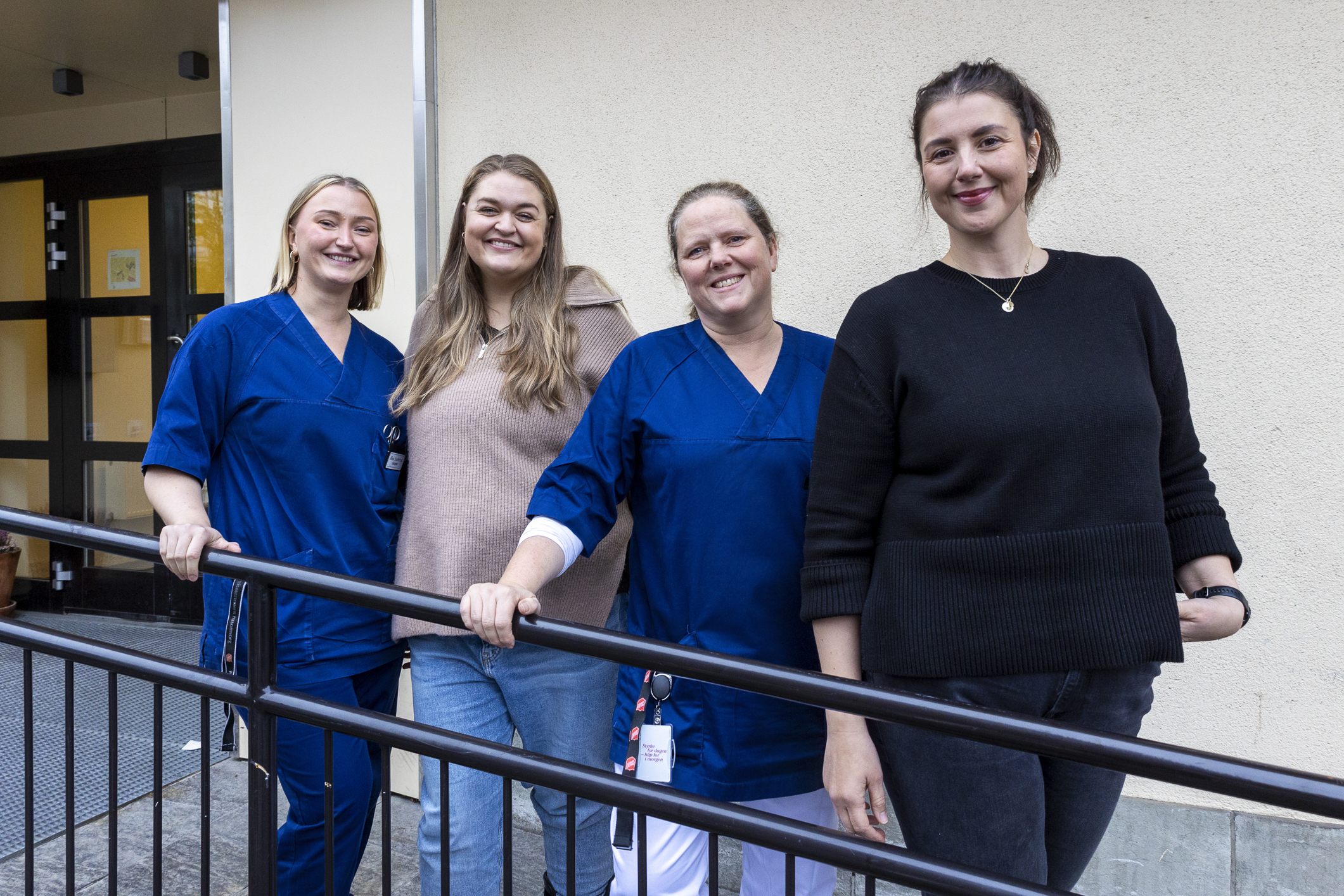 Fire kvinner står på rekke utenfor et murbygg. To av dem holder seg fast i rekkverket til rampen opp til inngangen.