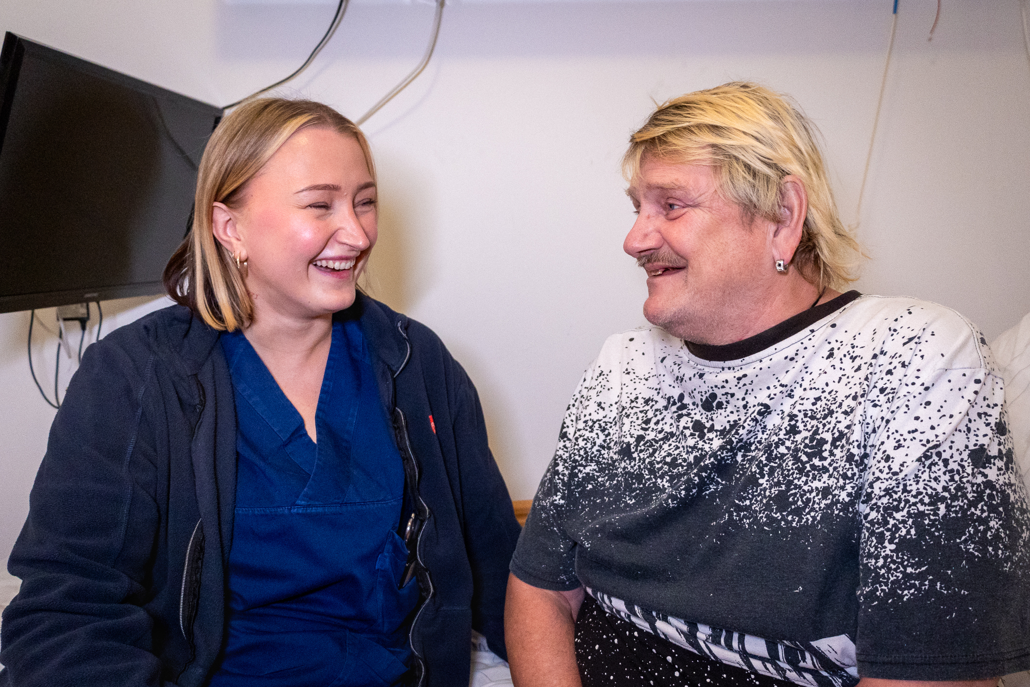 En kvinnelig sykepleier med blå uniform og en pasient som er en middelaldrende mann med bleket hår og bart, sitter på en sykeseng. De smiler til hverandre. En TV-skjerm er festet oppe på veggen til venstre over senga.