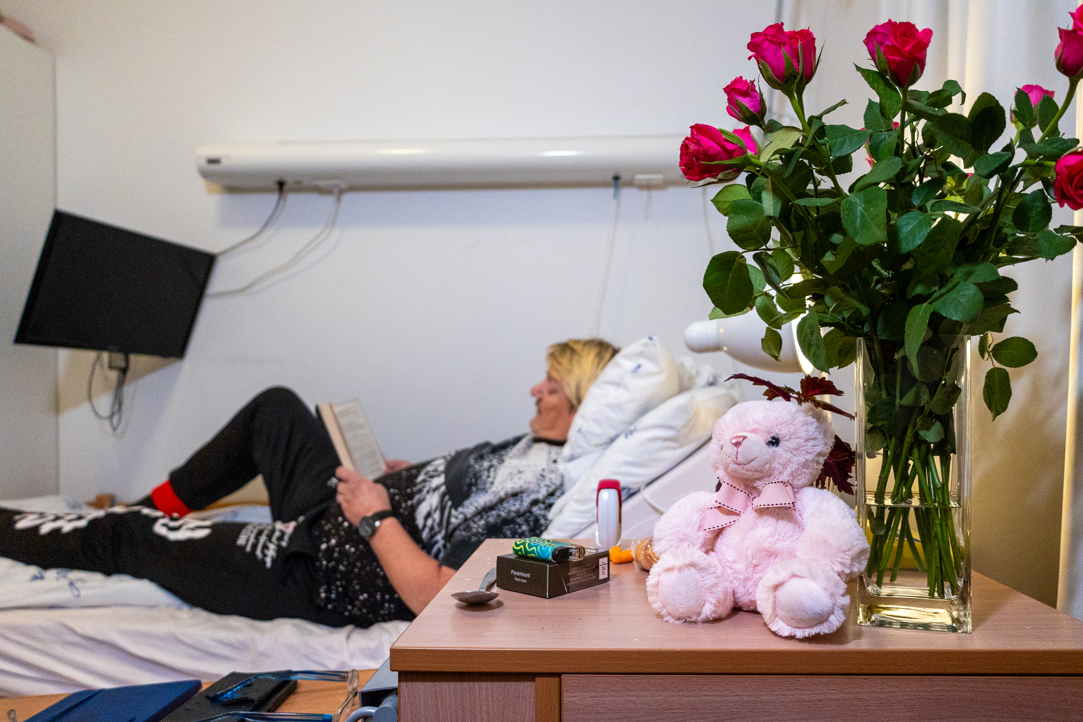 Middelaldrende mann ligger på en sykeseng og leser i en bok. På nattbordet står en rosa bamse og en vase med  røde roser