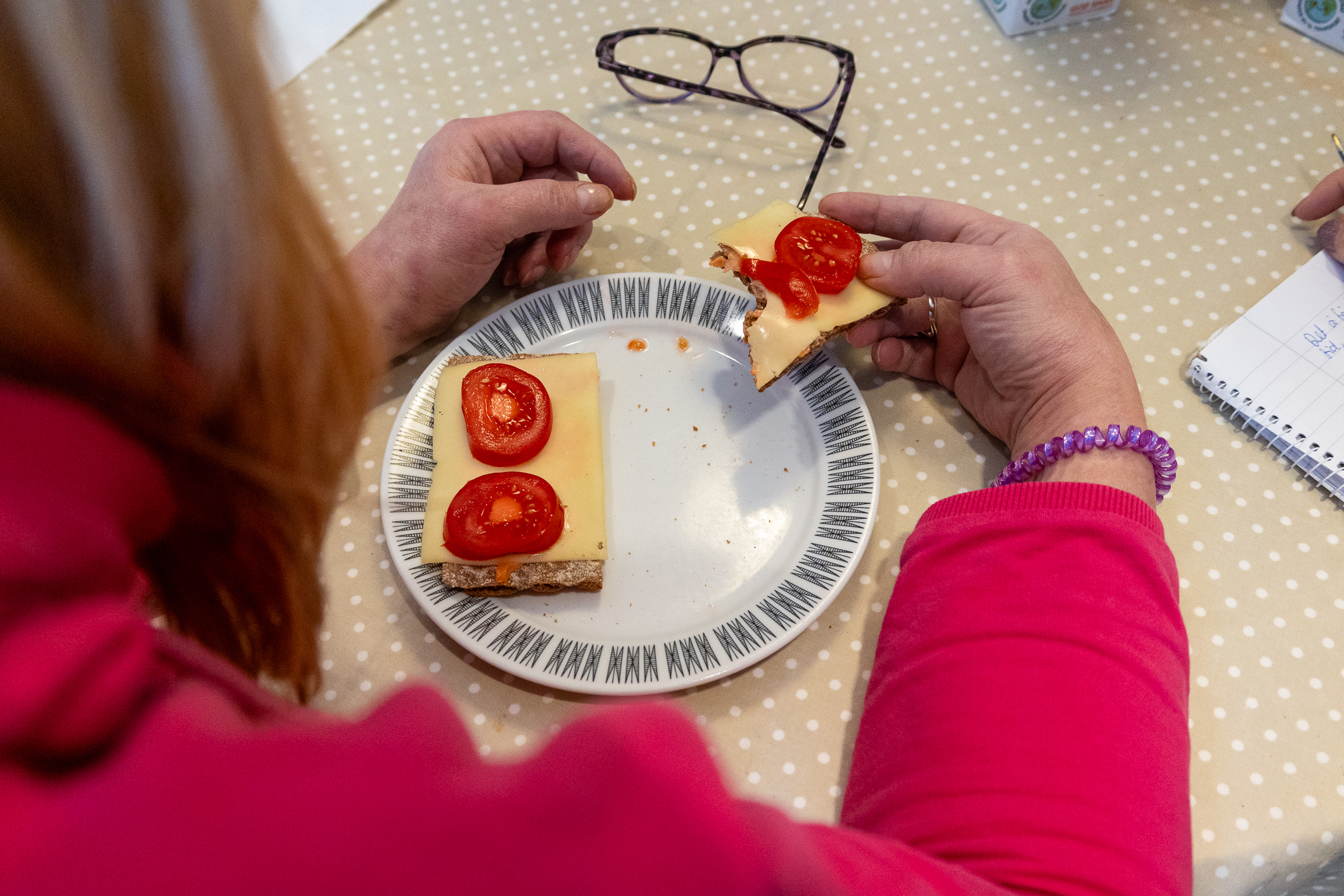 Kvinne i rød genser spiser ved et bord og spiser  brødskive med hvitost og tomat fra en tallerken