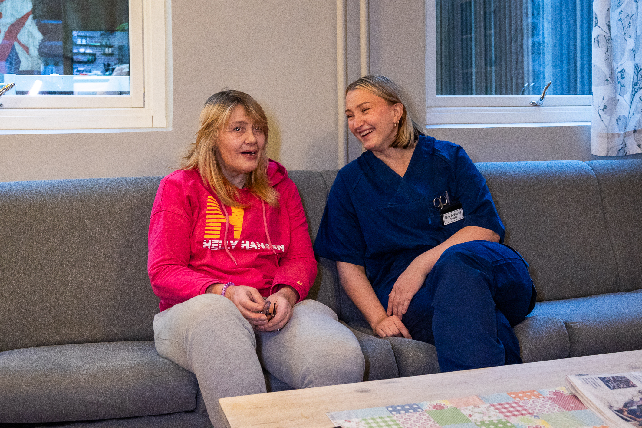 To middelaldrende kvinner sitter i sofaen. Den ene har rød genser og den andre har blå sykepleieruniform