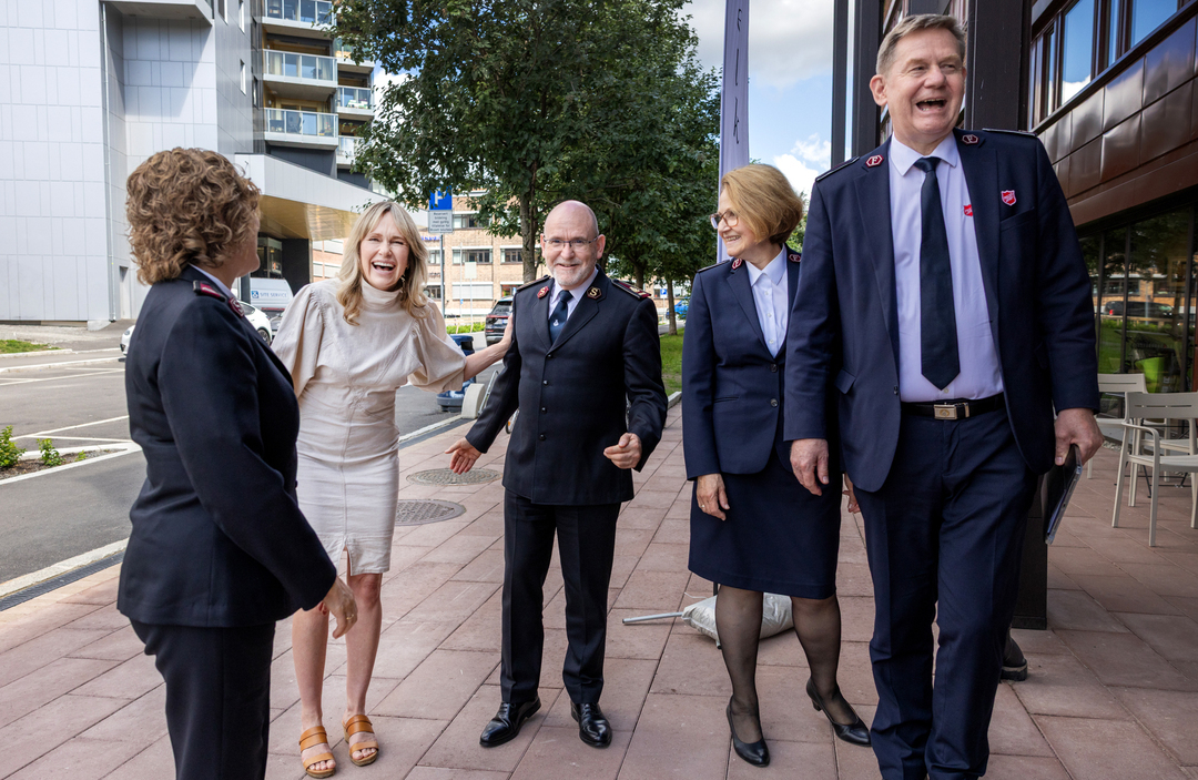 Fire voksne mennesker i uniform og én kvinne i lys kjole utenfor et bygg. De smiler og ler.