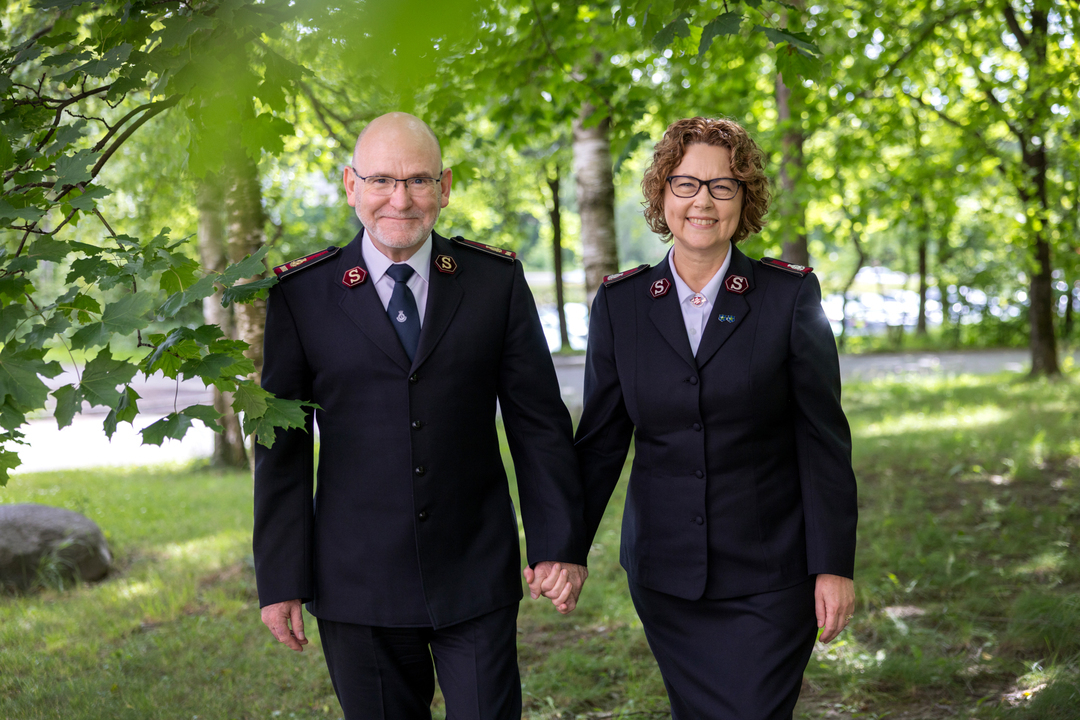 Et smilende, voksent par i mørk uniformer, hånd i hånd med grønne trær i bakgrunnen.