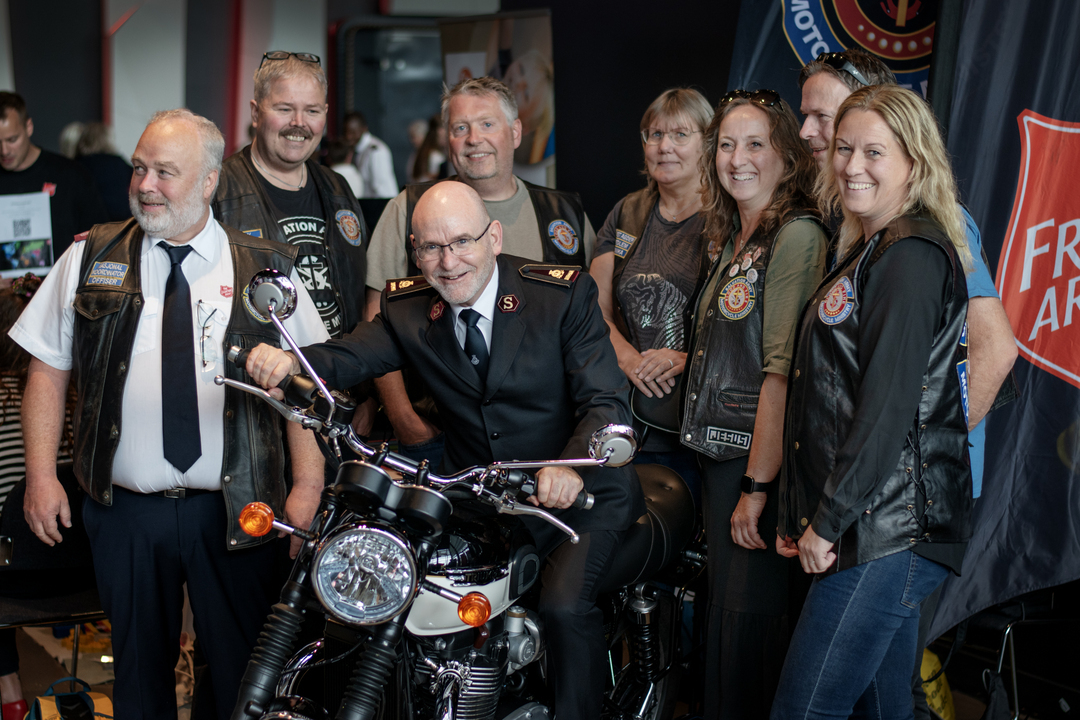 Foto: Middelaldrende, smilende, skallet mann med frelsesarméuniform på motorsykkel omgitt av en gjeng voksne motorsykkel-entusiaster.