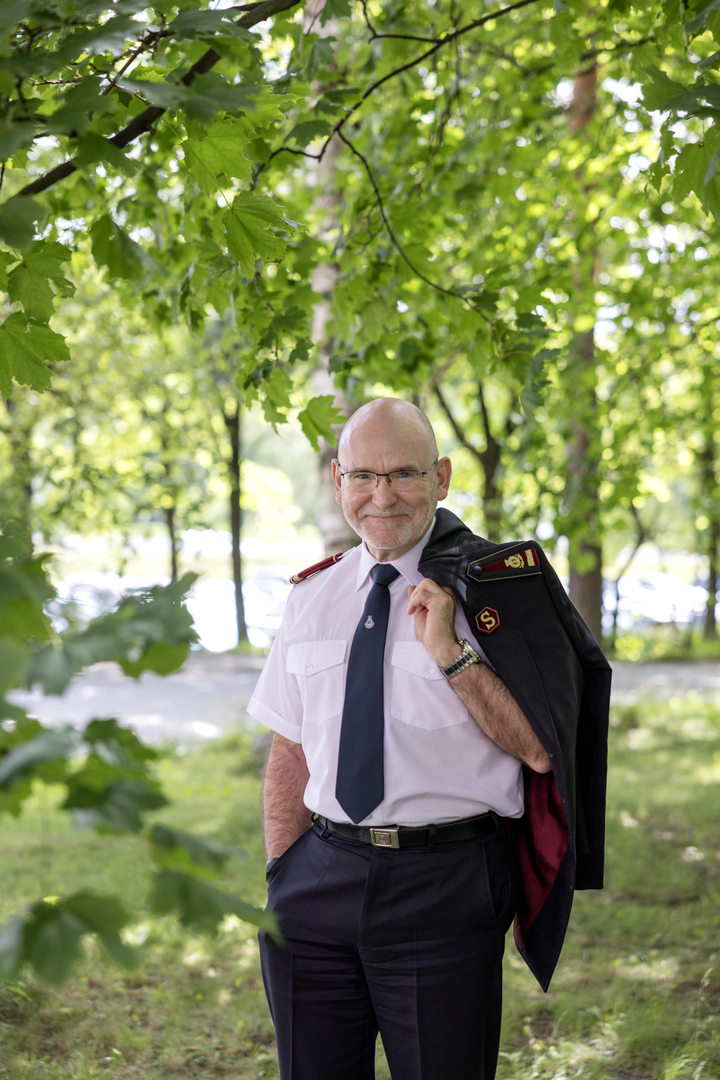 Portrettfoto av en smilende, skallet, middelaldrende mann med briller, hvit skjorte og sort frelsesarméuniformsjakke over skulderen. Bak ham er det grønne trær og gress.