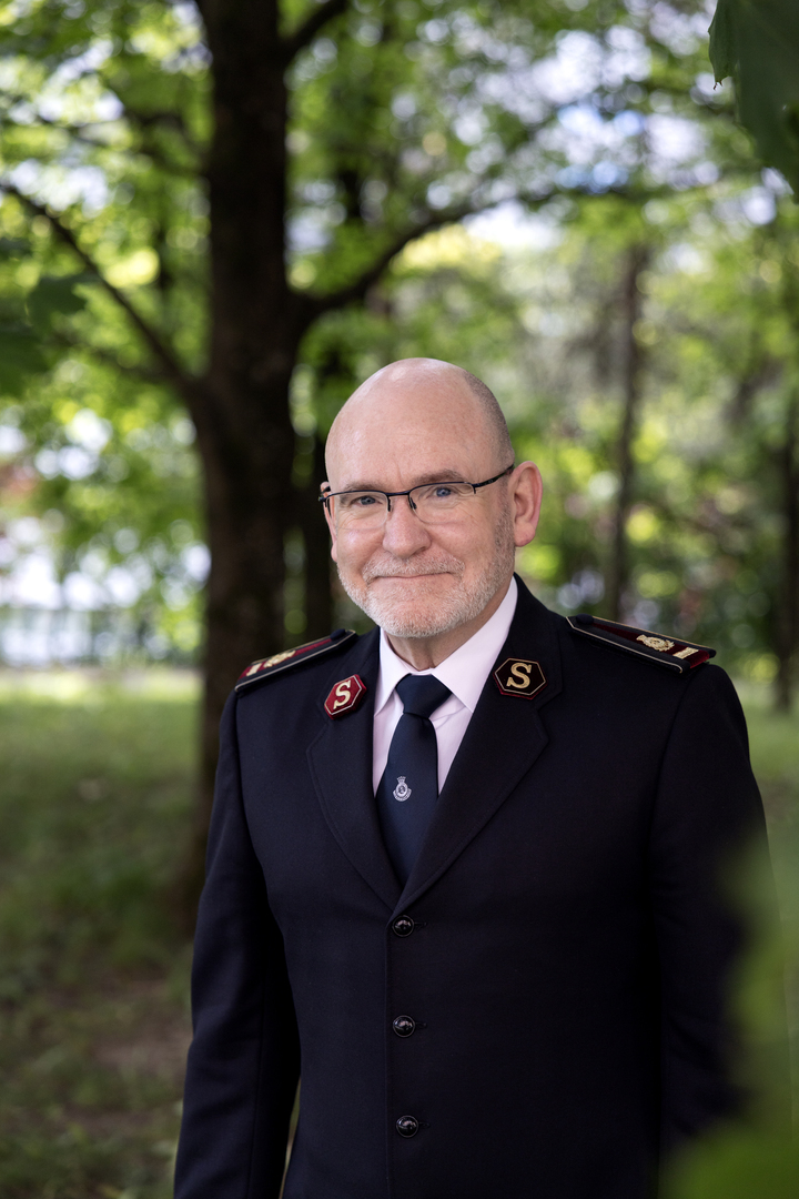 Portrettfoto av en smilende, skallet, middelaldrende mann med briller og sort frelsesarméuniform. Bak ham er det grønne trær og gress.