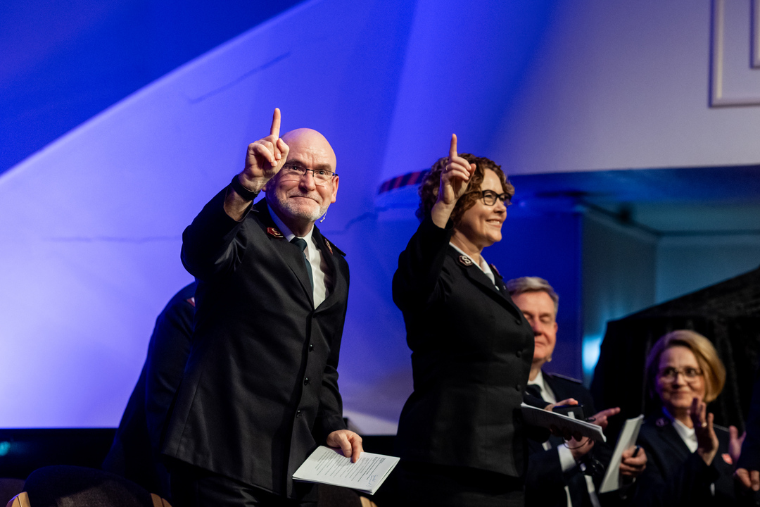 Et smilende, voksent par i mørk uniformer peker oppover. De står på en scene med blått lys.