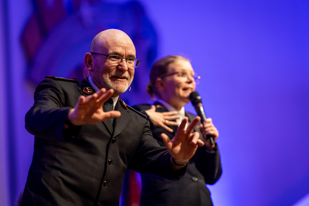 Foto: Skallet, middelaldrende mann med briller og sort frelsesarméuniform på scenen som gestikulerer og smiler med en yngre kvinnelig tolk bak med mikrofon. Hun har også uniform.