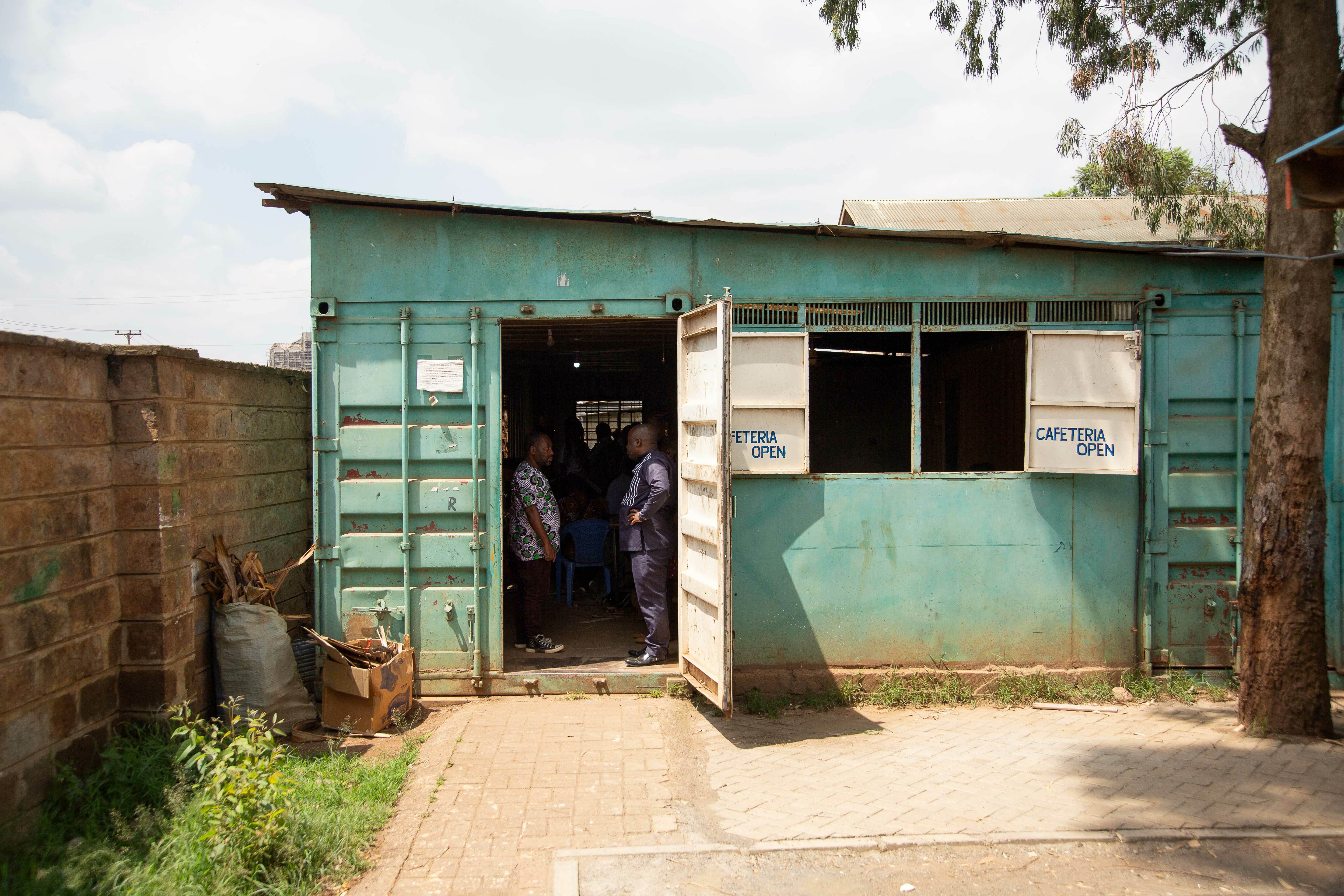 Grønt skur med et vindu og en åpen dør. I døråpningen står to afrikanske menn.
