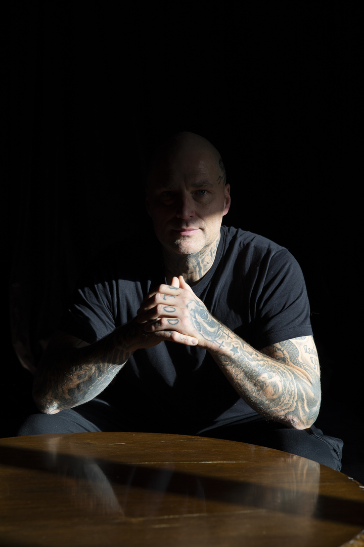 Mann med tatoveringer sitter ved et bord. På fingerne på den ene hånden står det BLOD. På armene ser vi tegninger av både Jesus og kors.