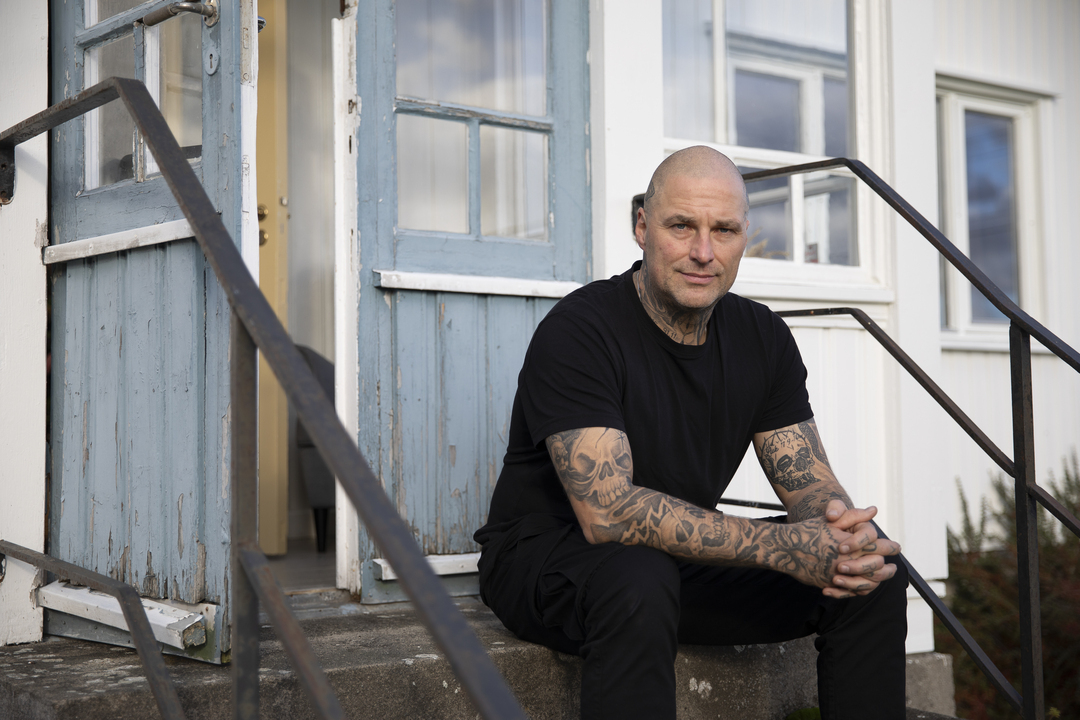 Mann med tatoveringer sitter på en trapp utenfor et hvitt hus med blå verandadør.