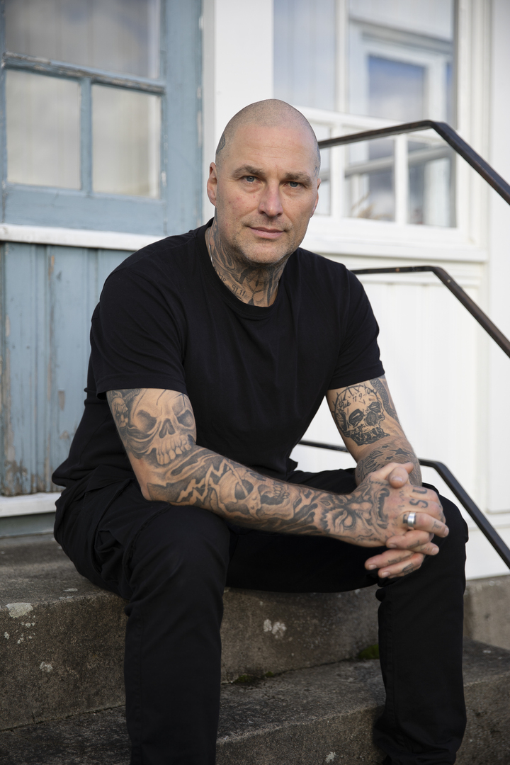 Mann med tatoveringer sitter på en trapp utenfor et hvitt hus med blå verandadør.