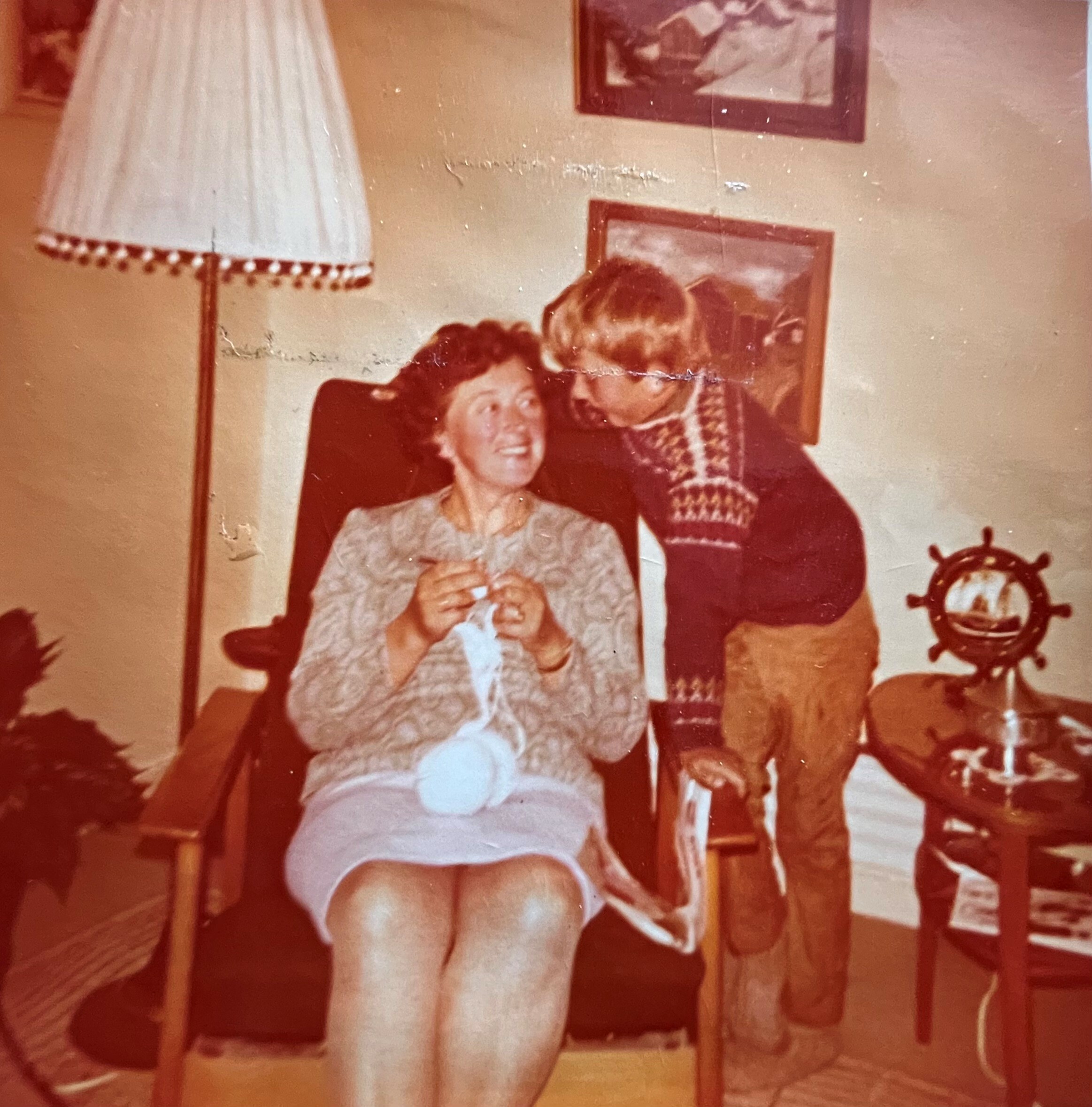 Gammelt bilde av mor som strikker og ung gutt som står ved siden av stolen hun sitter i.