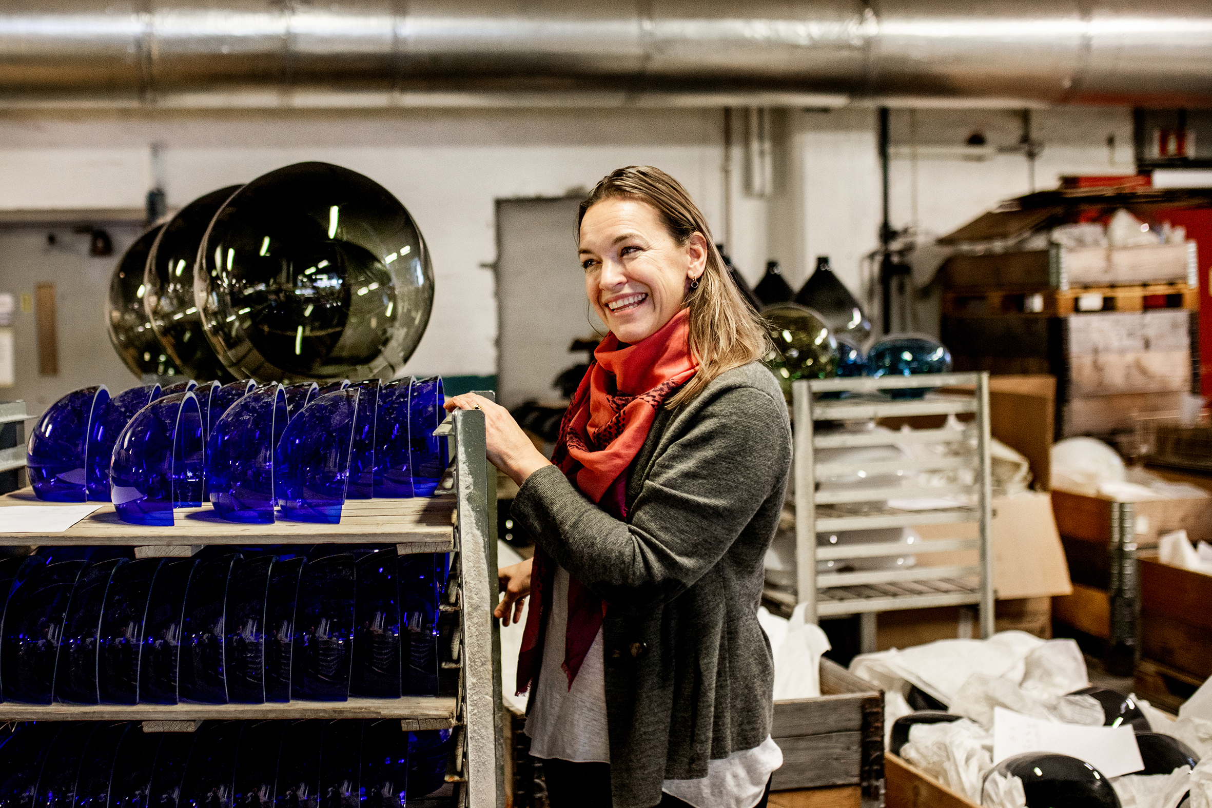 Bildet viser en kvinne med halvlangt hår, grå genser og rødt skjerf, som står foran en hylle med mange blå himmelvelvinger i glass som hører til julekrybber.