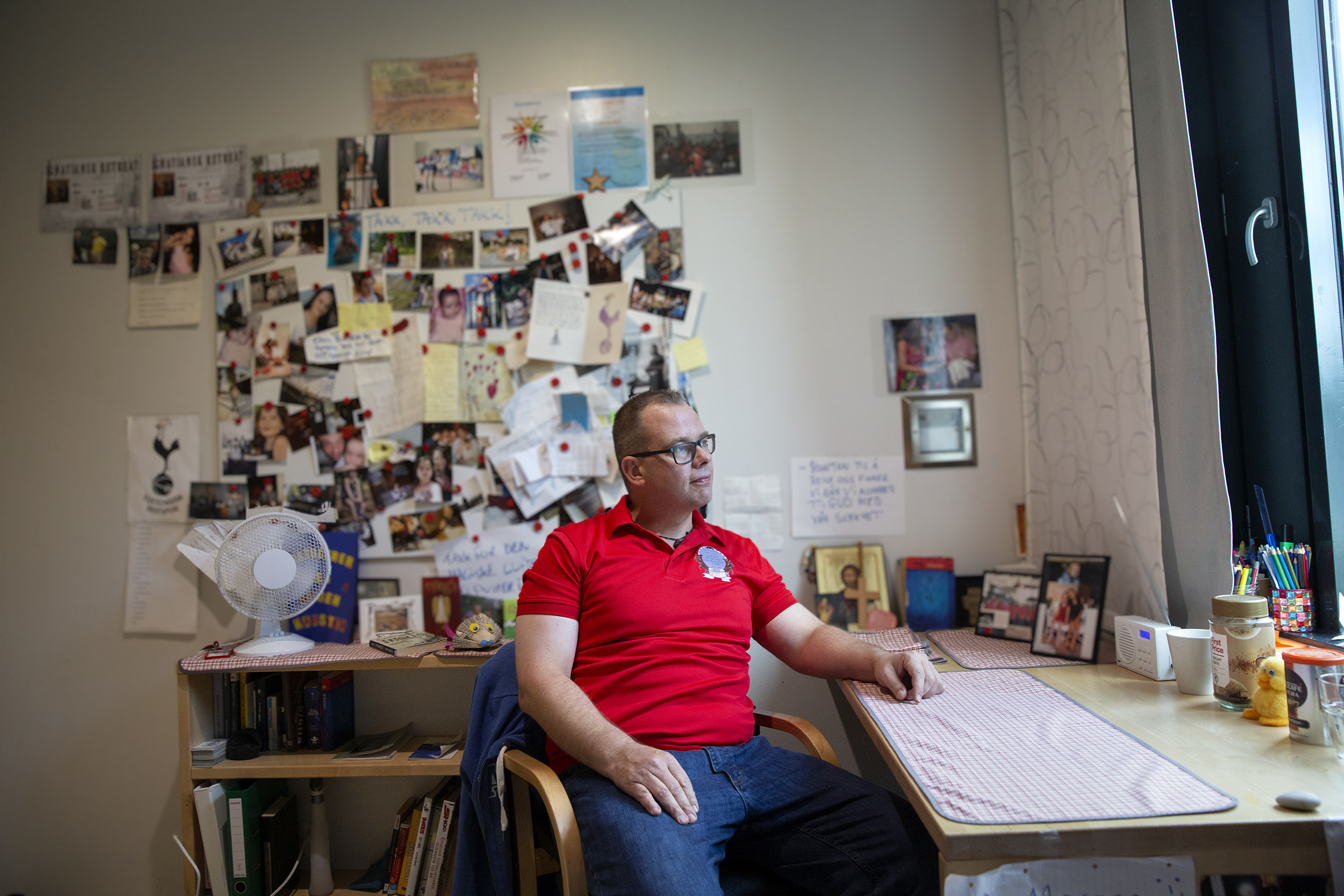 Carsten sitter på cellen sin, der er veggene dekorert med bilder og skrivebordet ryddig med et synlig Jesus ikon og et stort kors