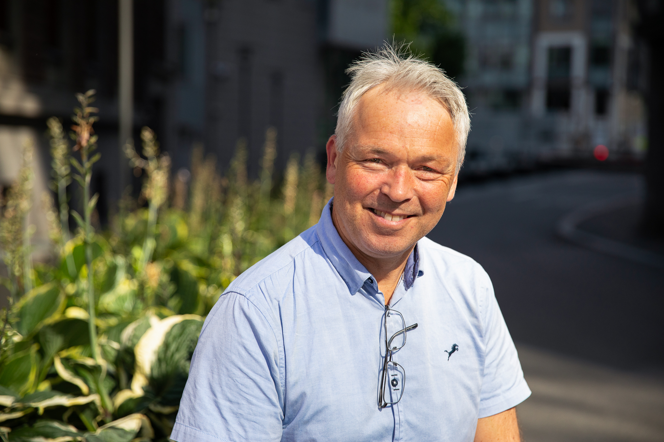 Solbrun mann i blå, kortermet skjorte smiler i sollyset utendørs.