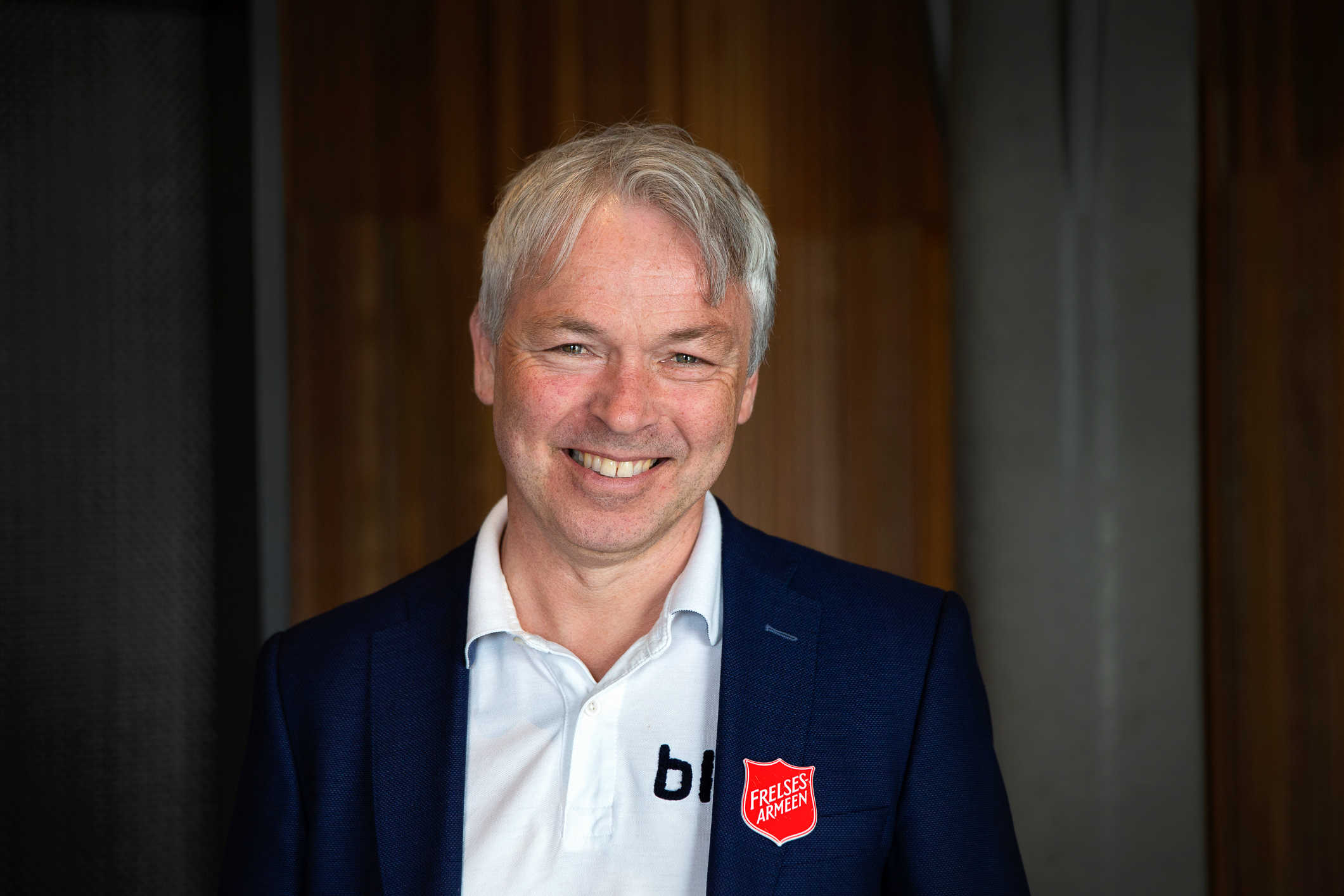Mann i hvit skjorte og blå dressjakke smiler mot kamera. På jakkeslaget har han en pin med logoen til Frelsesarmeen.