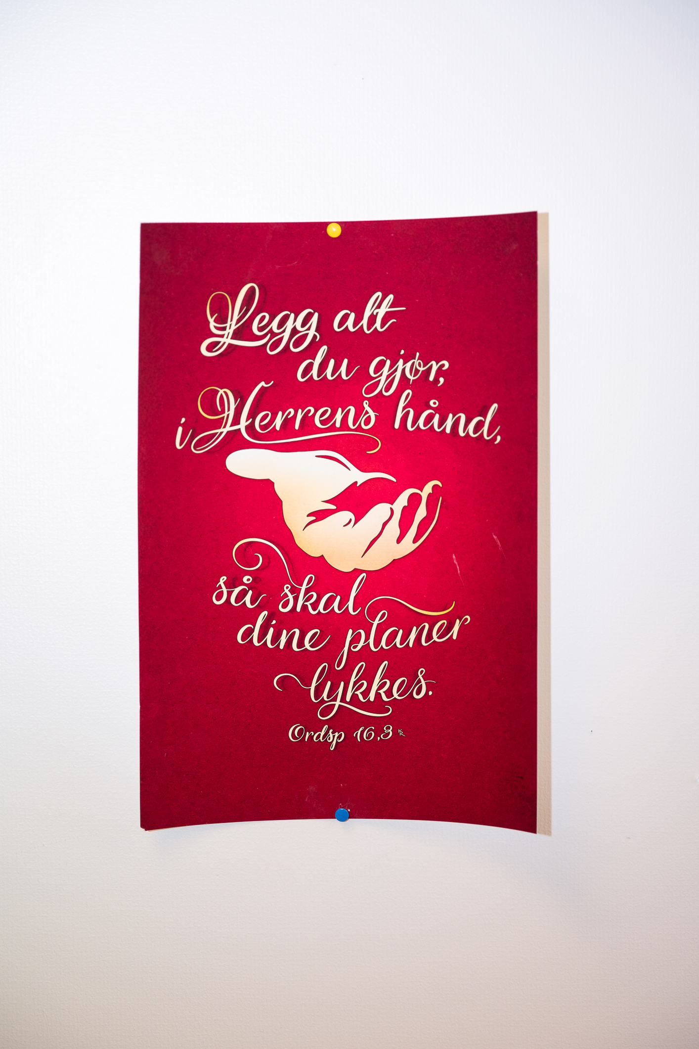 Bilde av et kort som henger på veggen der det står: "Legg alt du gjør i herrens hånd, så skal dine planer lykkes". Bibelverset er fra Ordspråkene 16,8.