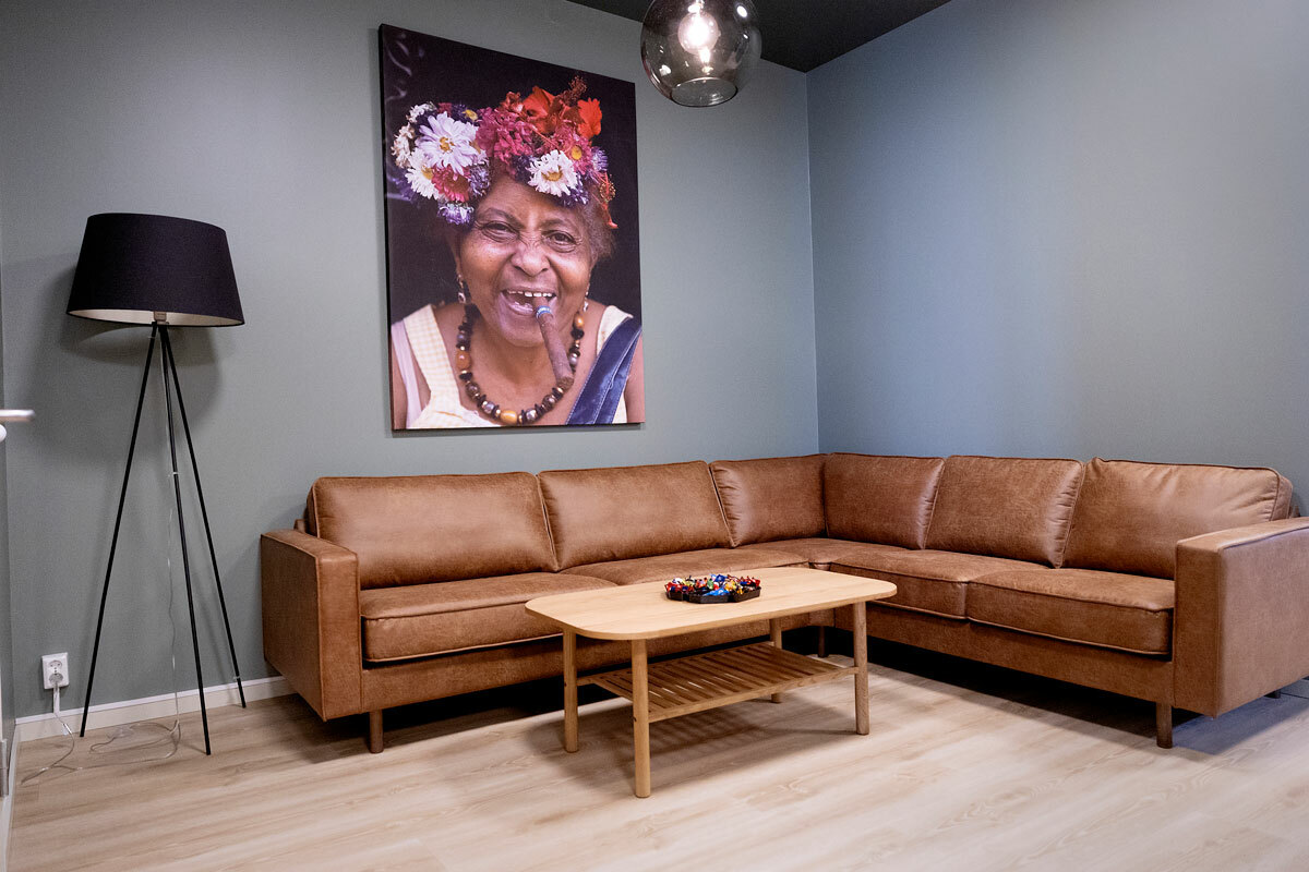 Foto: Del av en kro i en stue, men brun skinnsofa, bort, lampe og et stort fotografi på veggen av en gammel, cubansk dame med blomster i håret og en stor sigar i munnen.