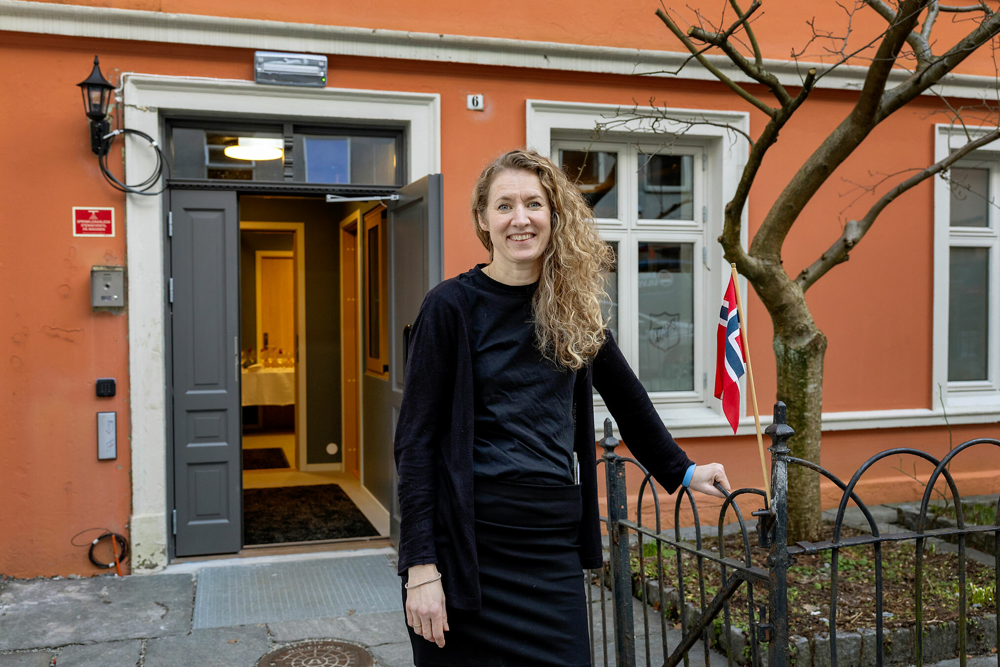 Foto: Kvinne i 40-årene med langt, krøllete hår smiler utenfor ei åpen dør inn i et murbygg.