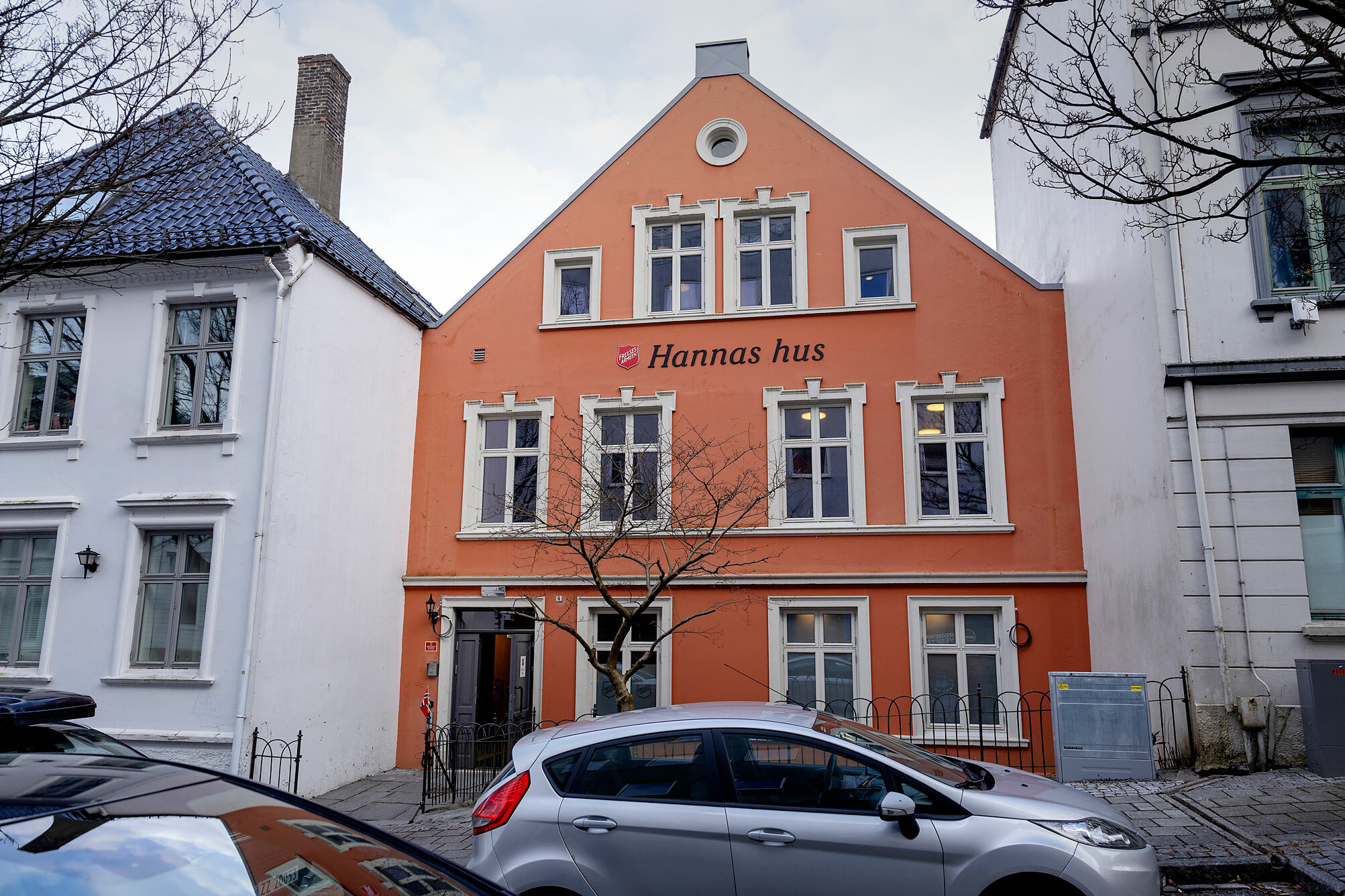 Foto: Gammelt, lakserosa murhus mellom to andre, hvite hus. På fasaden, mellom vinduene i andre og tredje etasje står det Hannas hus bak Frelsesarmeens røde logo.