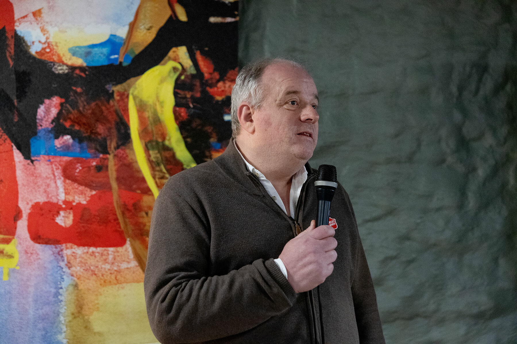 Foto: Mann i 60-årene med grått hår snakker i en mikrofon. Bak ham henger et fargerikt bilde.
