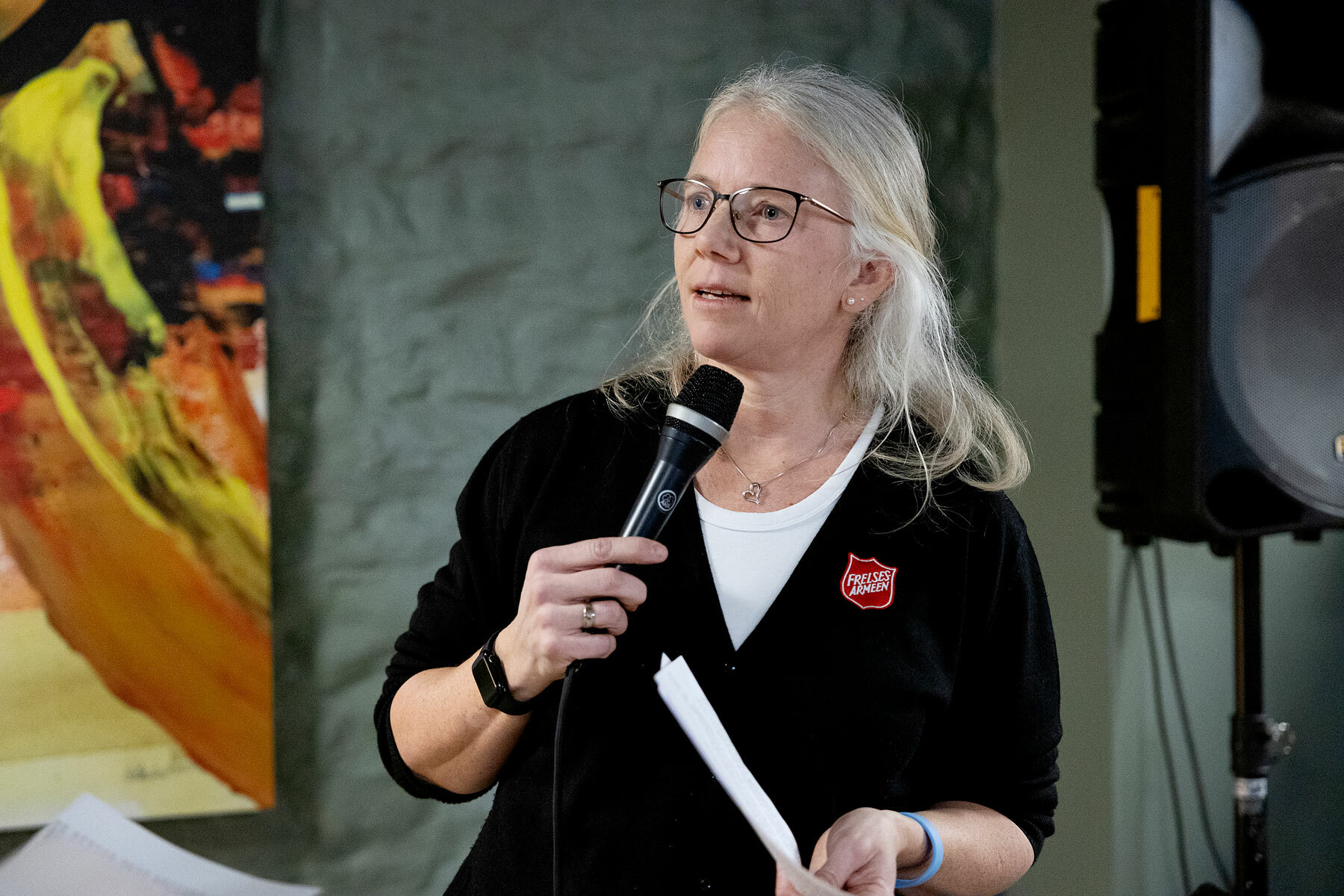 Foto: Kvinne med langt, gråblondt hår og briller snakker i mikrofon. Hun har Frelsesarmeens røde logo på jakken.
