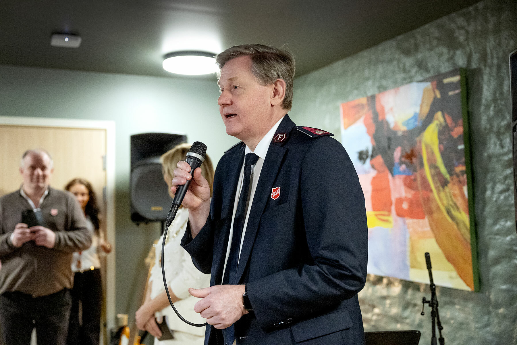 Foto: Mann i 60-årene med uniform fra Frelsesarmeen snakker i en mikrofon. På høyre side av ham står det flere og lytter.