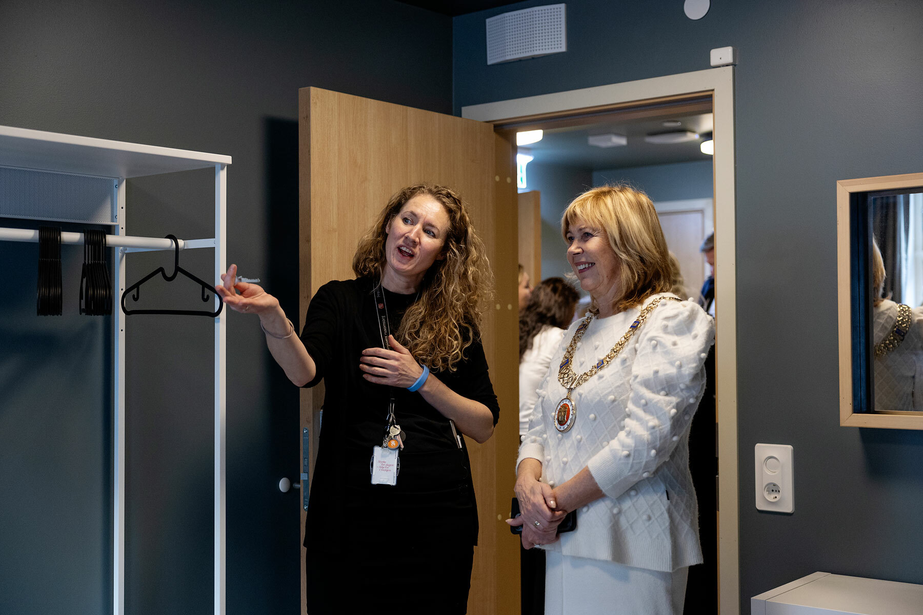 Foto: Kvinne med langt, krøllete hår peker innover i et rom. I dørkarmen står en dame i hvit drakt med ordførerkjede rundt halsen.