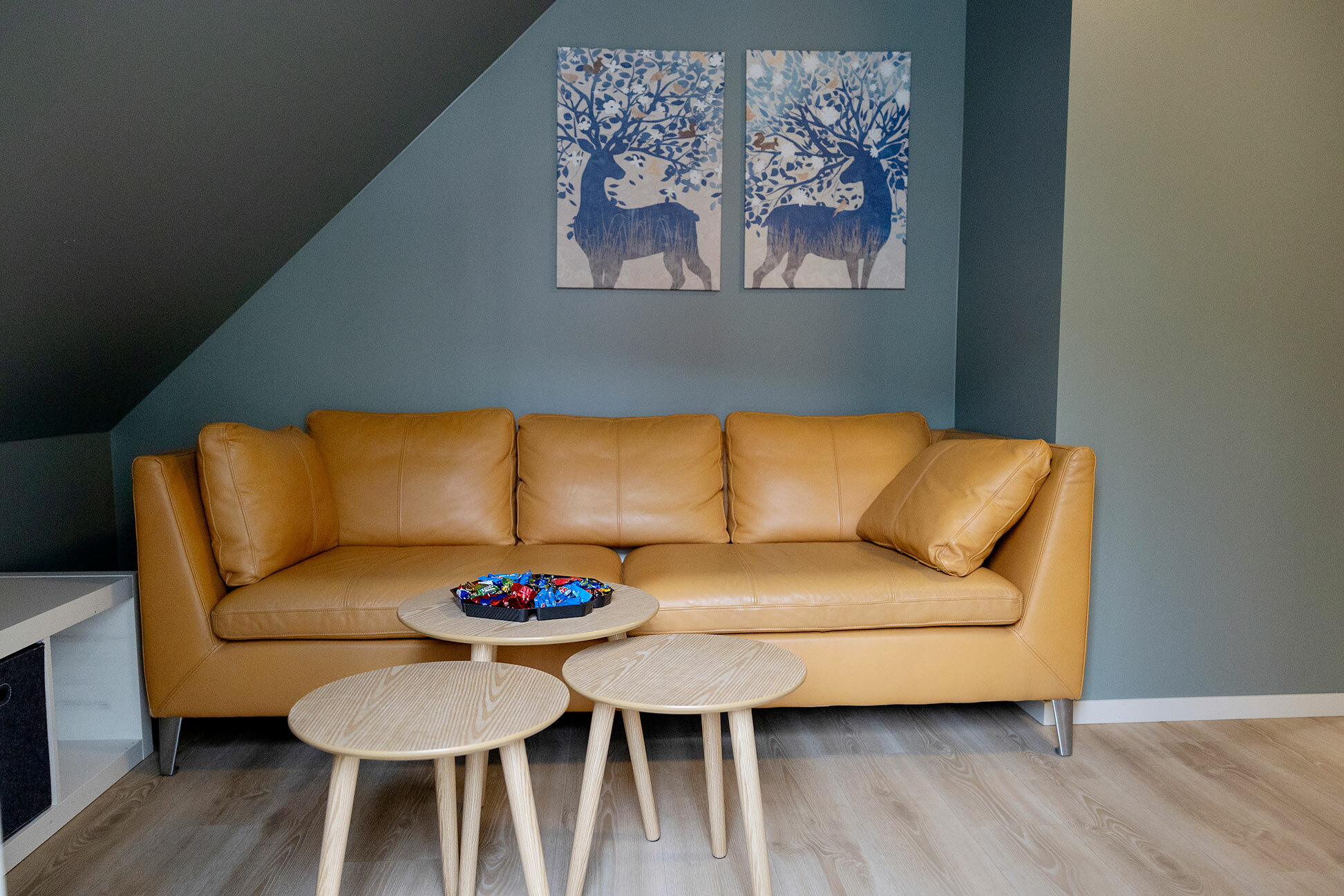 Foto: Av en gul skinnsofa under et skråtak. Fargen på veggene er grønngrå og det henger to kunstverk på veggen av hjortedyr.
