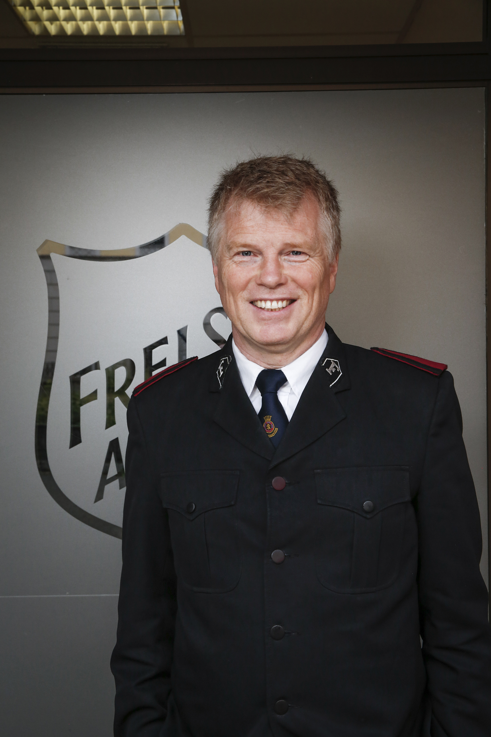 Ola Grytten, økonomiprofessor, historiker og soldat i Frelsesarmeen. Her i Frelsesarmeen uniform foran en grå vegg med Frelsesarmeen skjoldet (Foto: Mette Randem