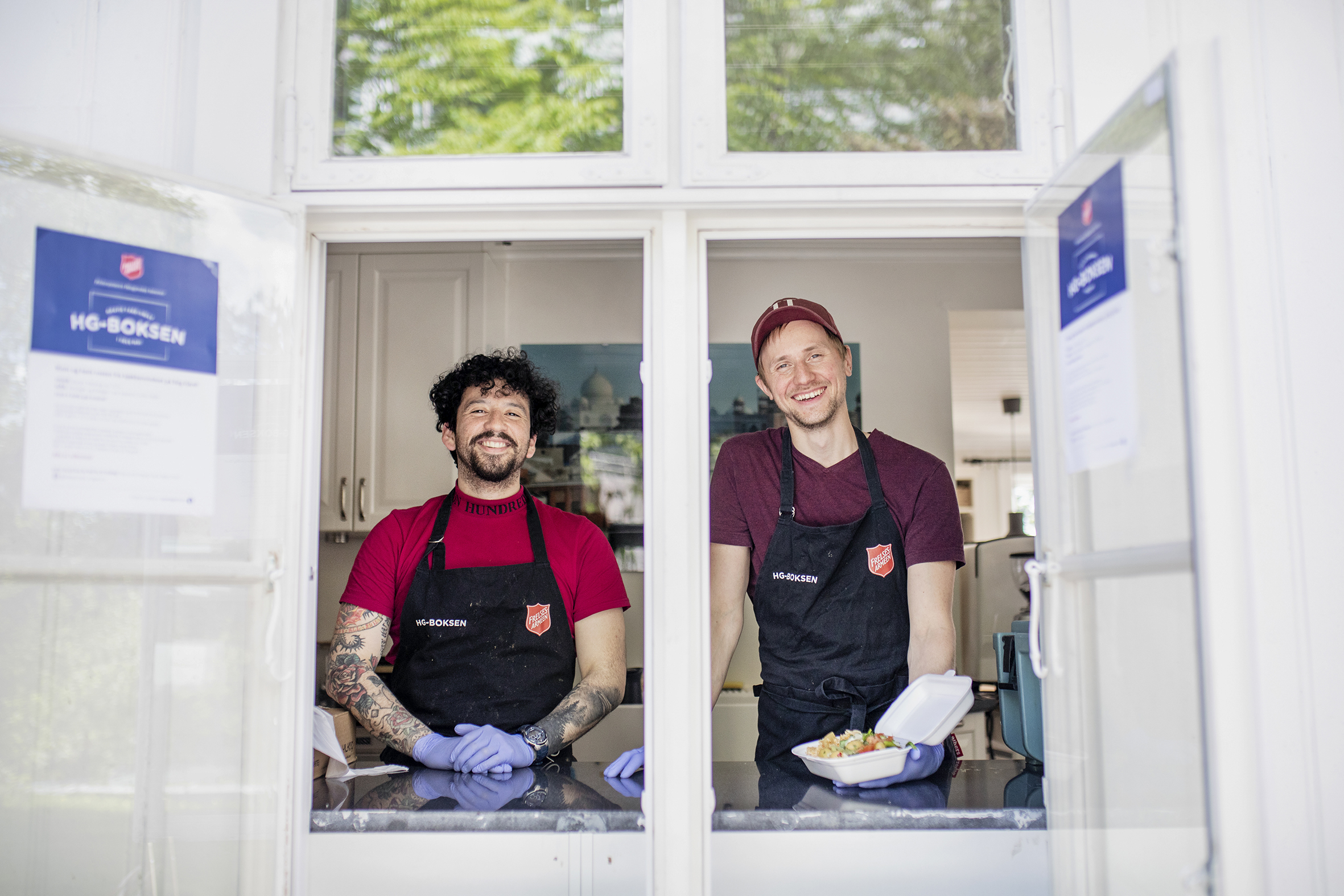 Felipe Castro (t.v) og Andreas Welander (t.h)  står i kjøkkenviduet hvor de deler ut mat. De har på seg forklær med Frelsesarmeens logo. Andreas holder en åpen boks med mat i hånda.