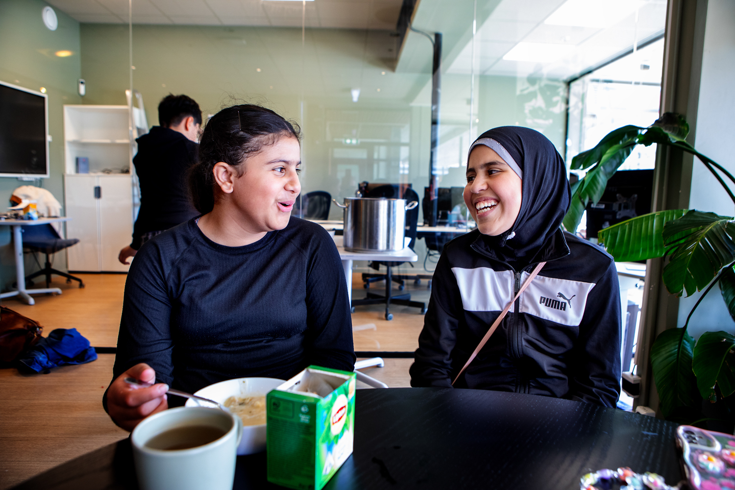 Foto: To jenter sitter og smiler mot hverandre. Rand til venstre har på seg svart genser, hun holder en skje og skal til å spise. Tabarek til høyre har på seg svart hijab og svart og hvit treningsjakke.