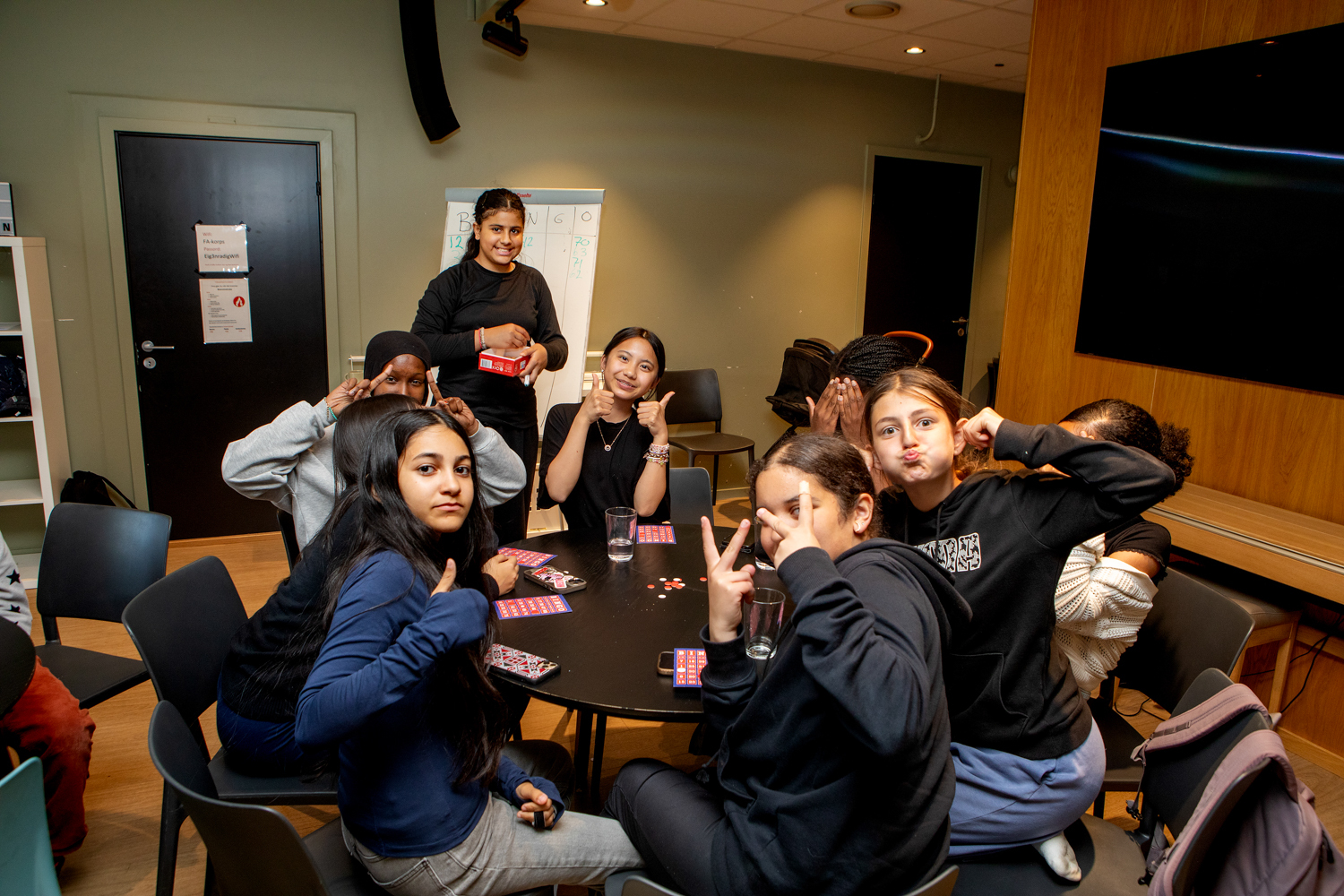 Foto: En gruppe jenter sitter rundt et rundt bord og smiler, flere av dem viser tommel opp eller gjør grimaser. Bak dem står Rand og holder bingospillet.