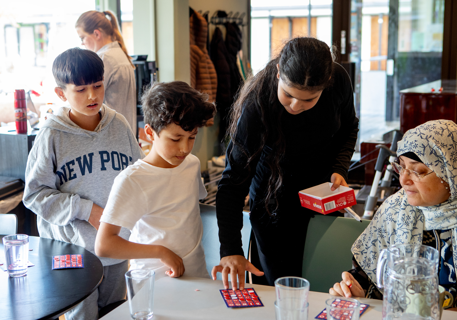 Foto: Rand står i midten av bildet med svart langt hår og svarte klær. Hun bøyer seg over et bord og sjekker et bingobrett. To gutter står til venstre og følger med, til høyre sitter en kvinne med sjal over håret og ser på.