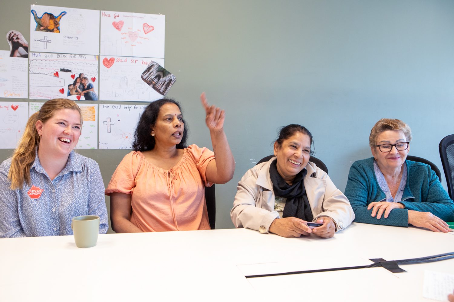 Foto: Åse Kari i blå skjorte med Frelsesarme-skjold sitter ved et bord ved siden av tre kvinner. Alle smiler og ser mot siden.