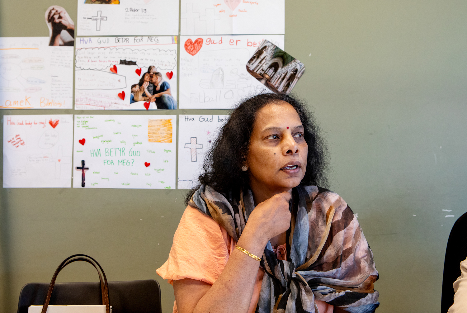 Foto: En kvinne med langt mørkt hår, fersken farget bluse og mønstrete sjal sitter ved et bord og ser mot siden. På veggen bak henne er meninger og plakater laget av barn.