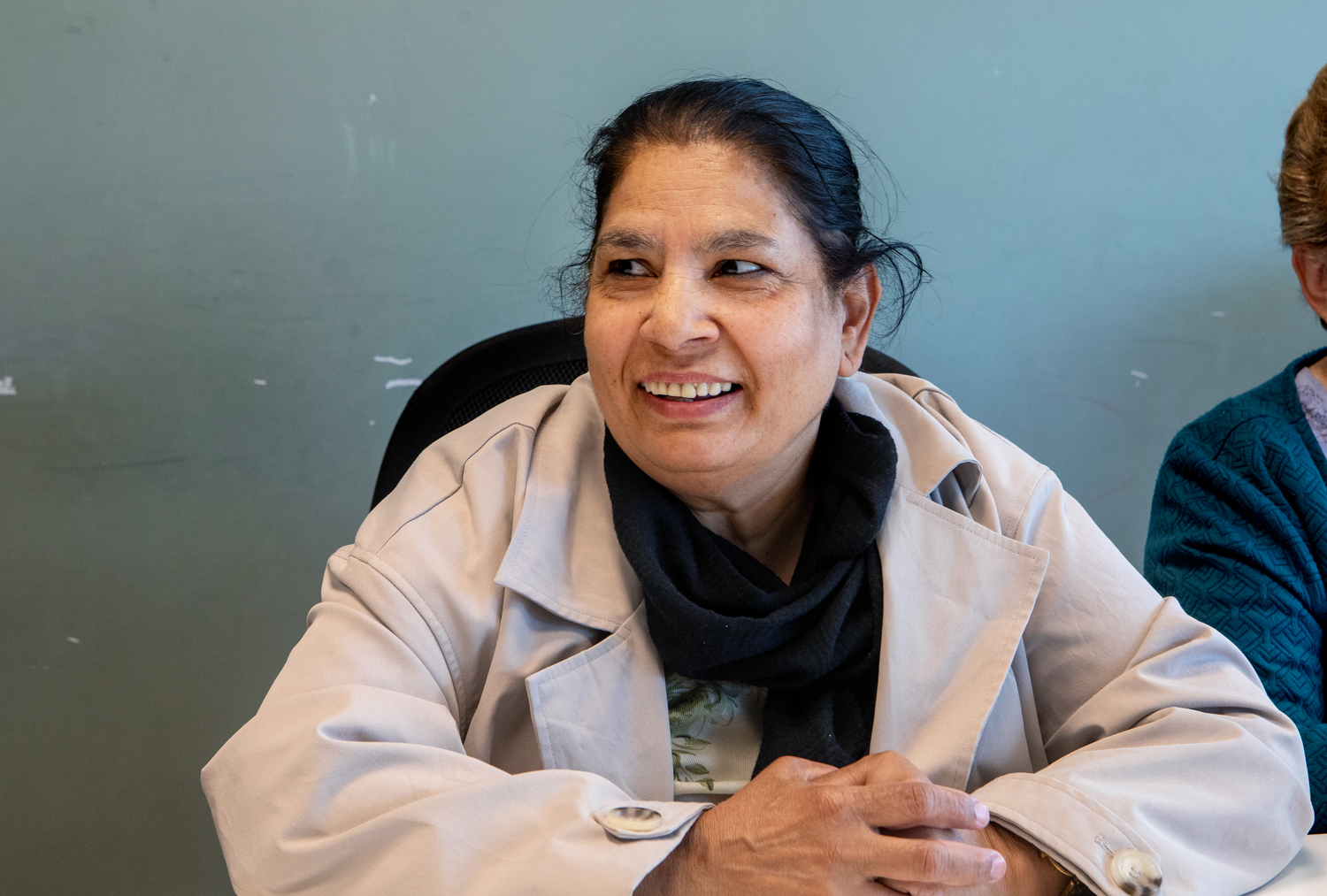 Foto: En kvinne med mørkt langt hår dratt bakover og beige jakke sitter og smiler og ser mot siden.