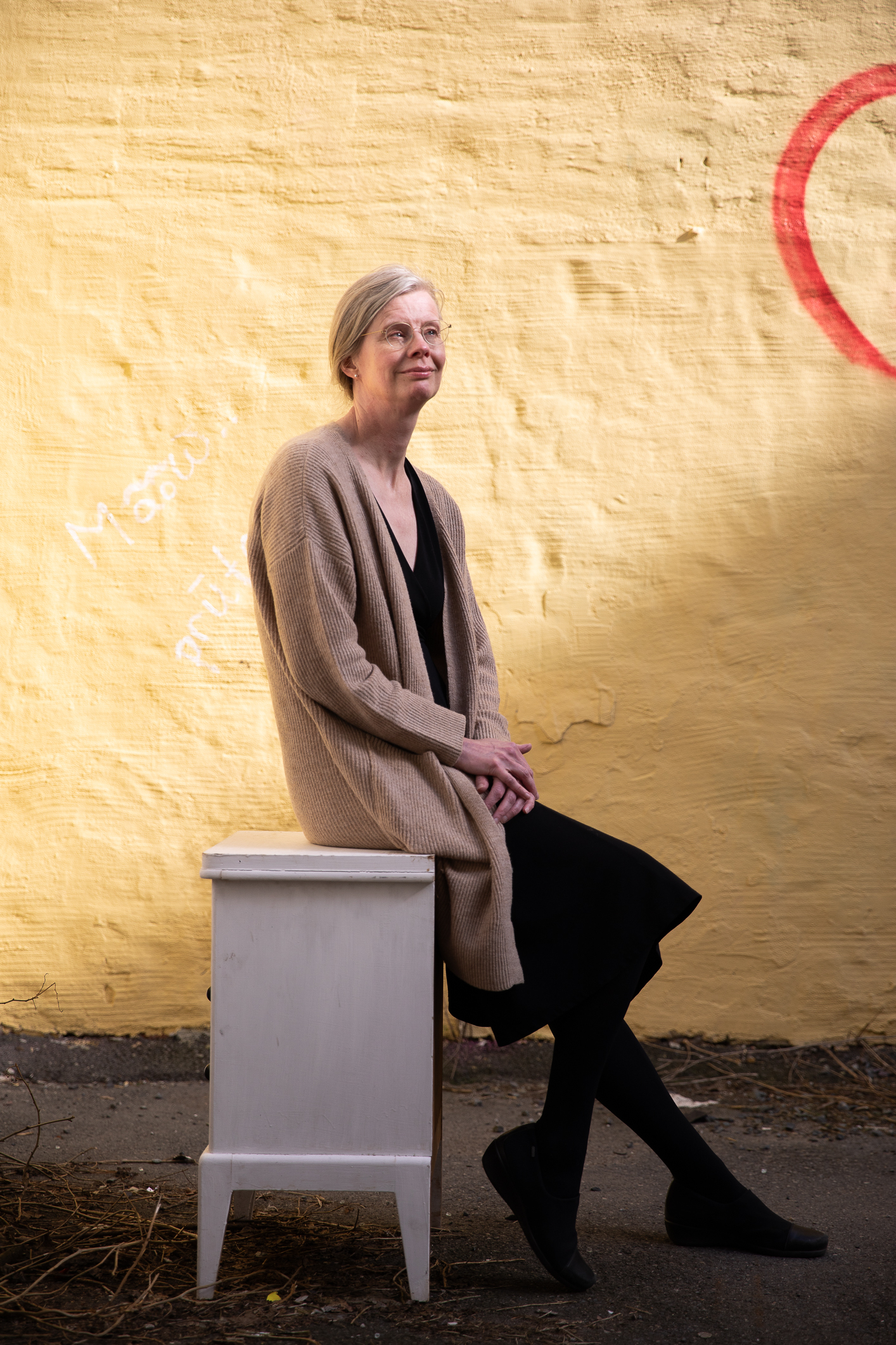 Dame, Hedvig Montgomery, sitter lent inntil et lite nattbord med en gul murvegg i bakgrunnen.