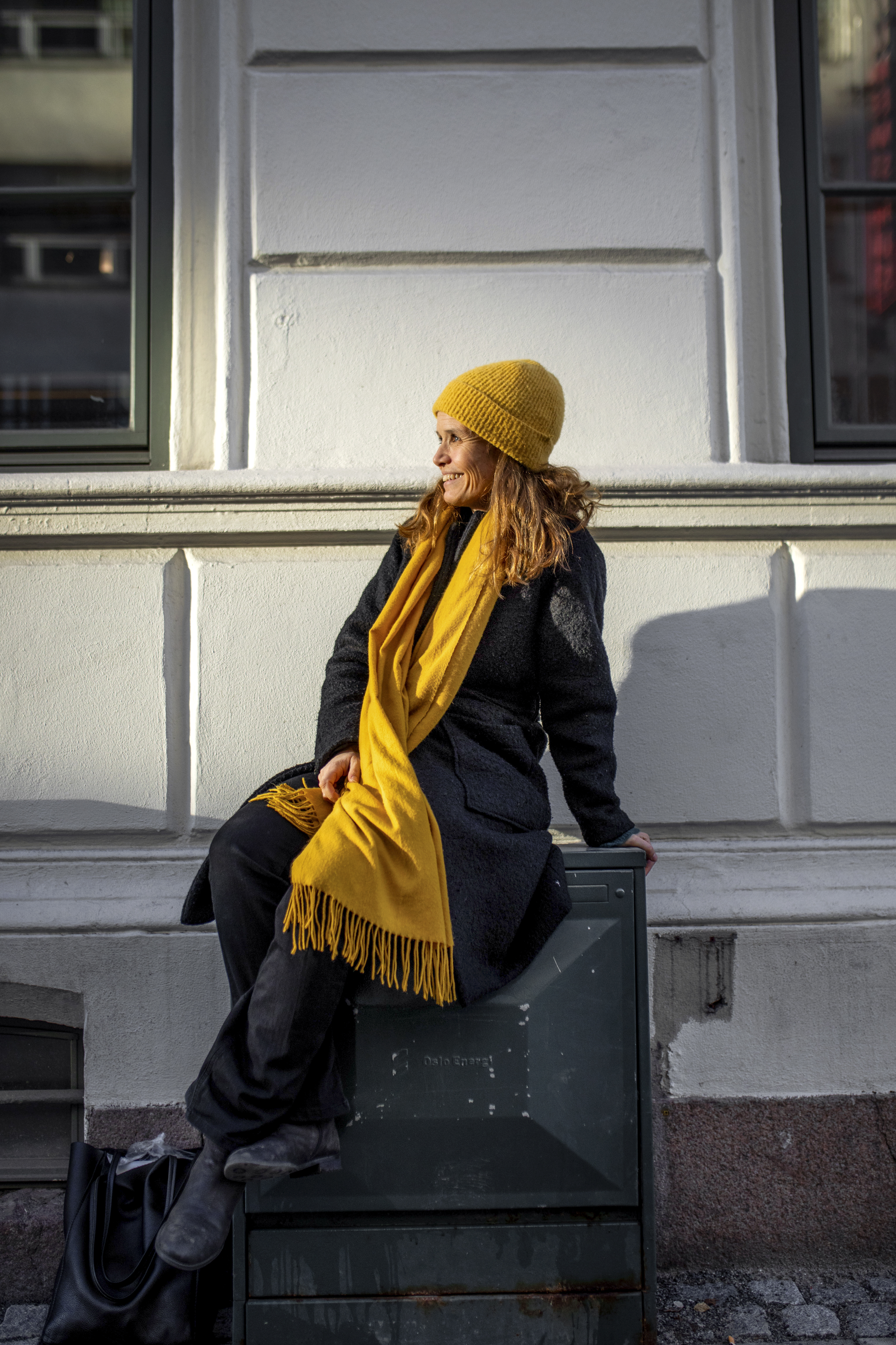 Helga Samset sitter på en boks mot en murvegg i Oslo sentrum. Hun smiler mot sollyset som treffer henne .