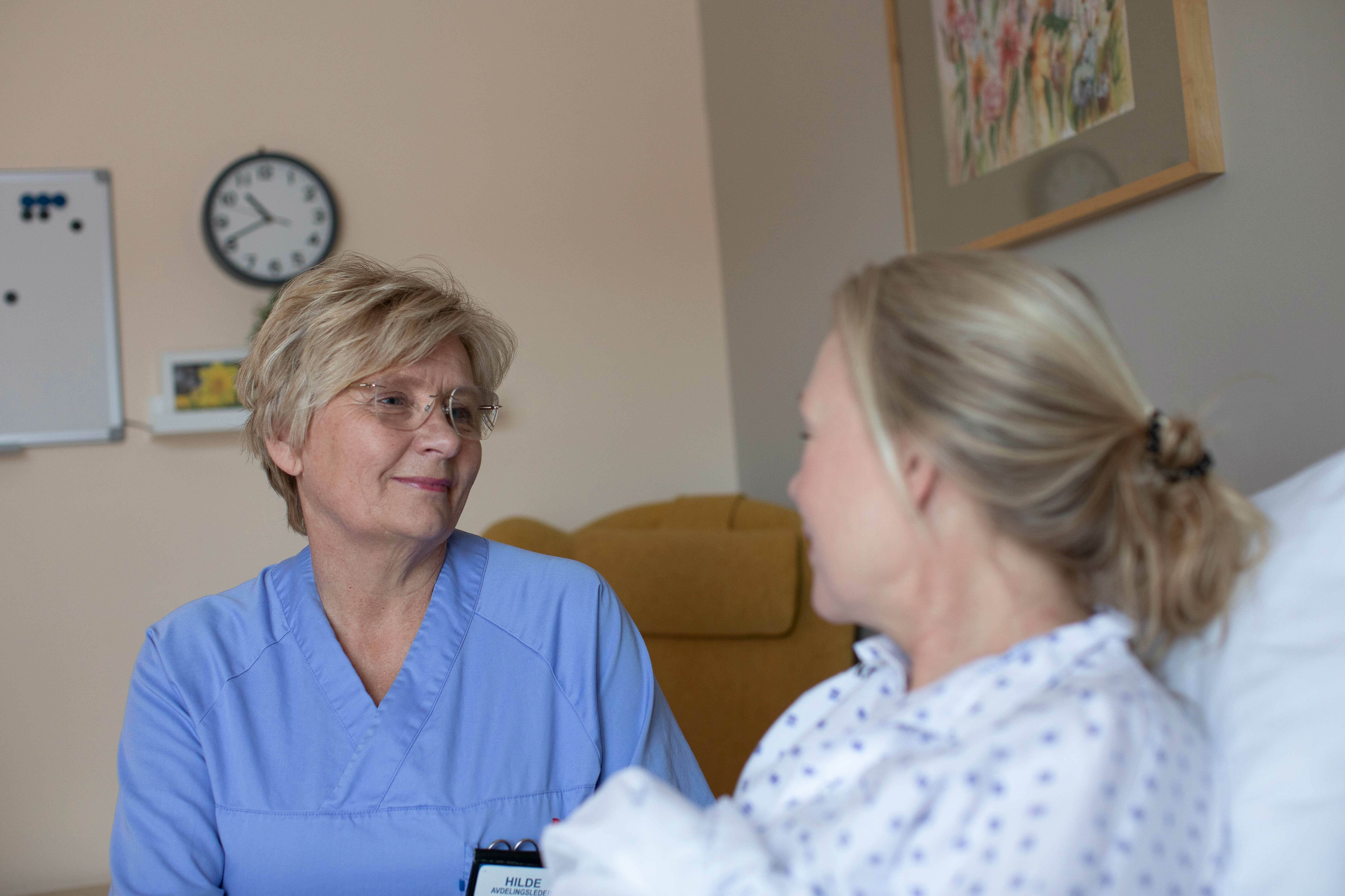 En sykepleier i blå uniform sitter og snakker med en pasientent som er en ung kvinne.