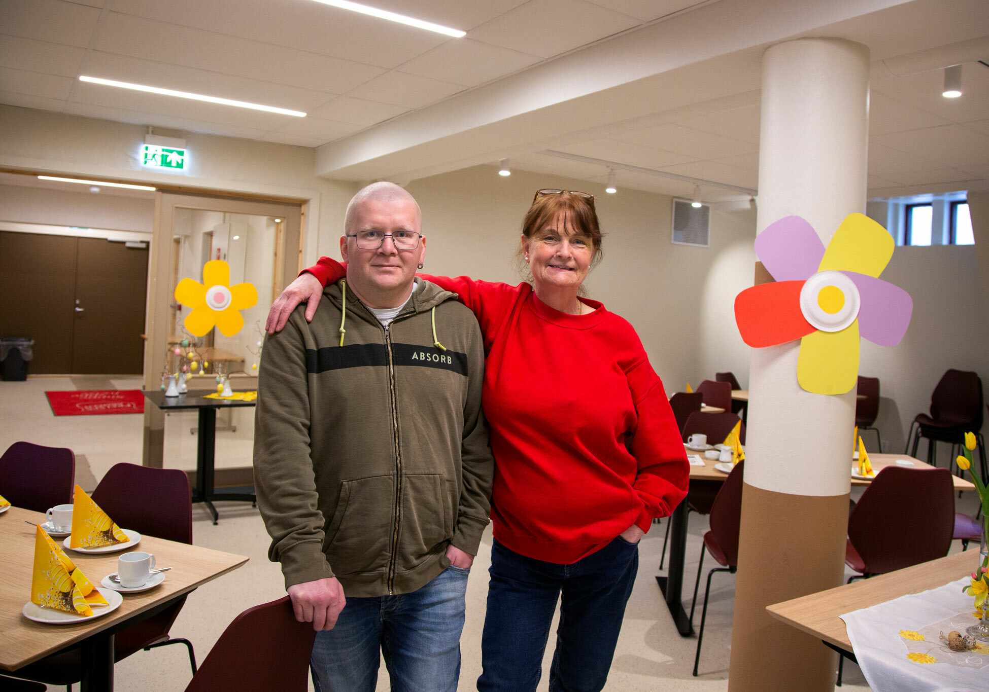 Foto: Midt i et rom med dekkede bord står en mann og en kvinne midt i livet. Han har på seg grønn collegejakke med hette. Hun holder armen sin rundt ham, og har på seg en knall rød genser. På en søyle og veggen bak er det laget store, fargerike blomster av papir.